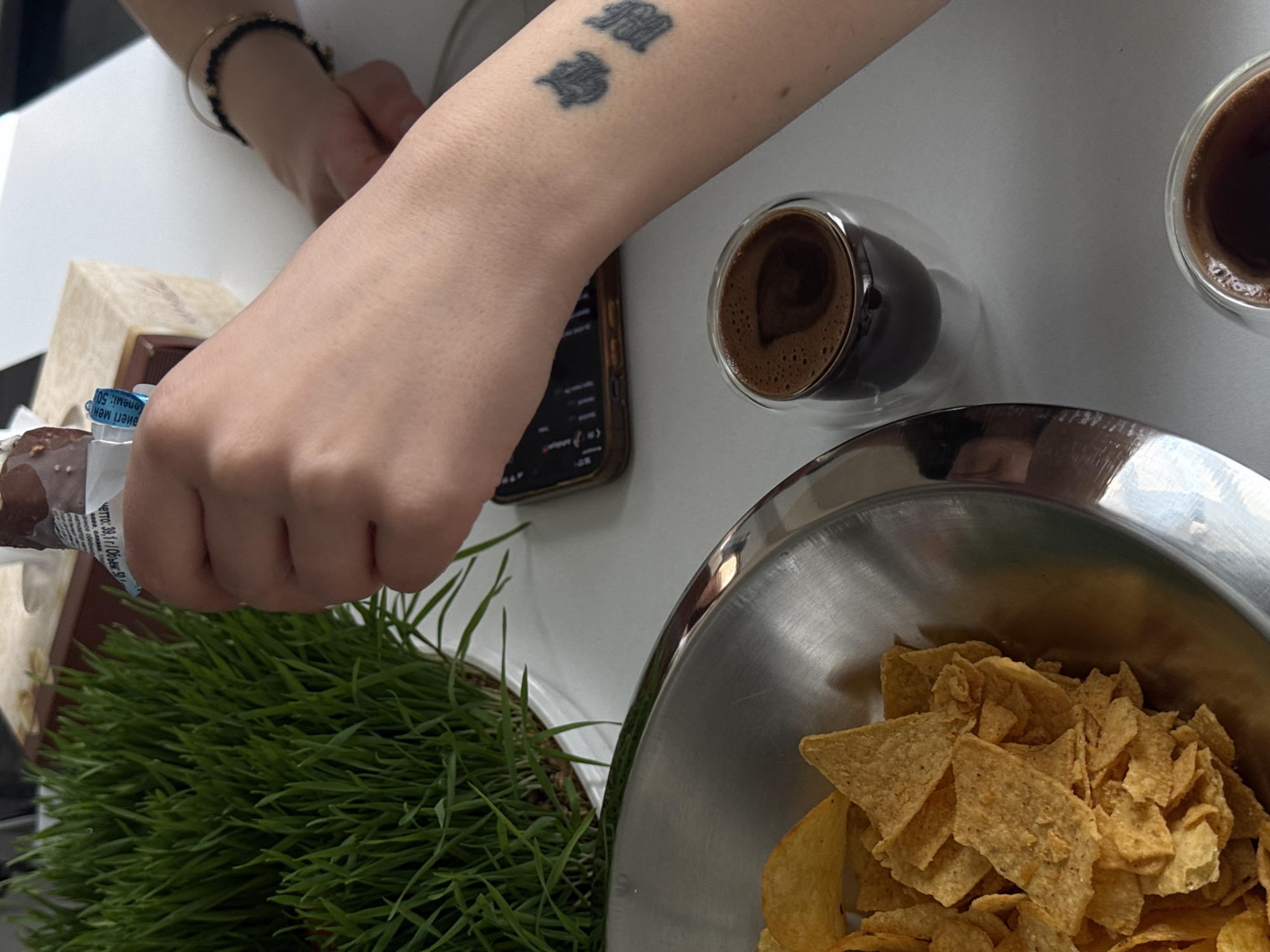 A top-down shot shows a person's tattooed arm near a glass of coffee, a bowl of tortilla chips, and a container with sprouts. The person is holding the container of sprouts. Part of a phone is visible under the arm, and a second coffee is placed at the top right of the frame. The product label contains text in Russian or a related language. Location information is unavailable.