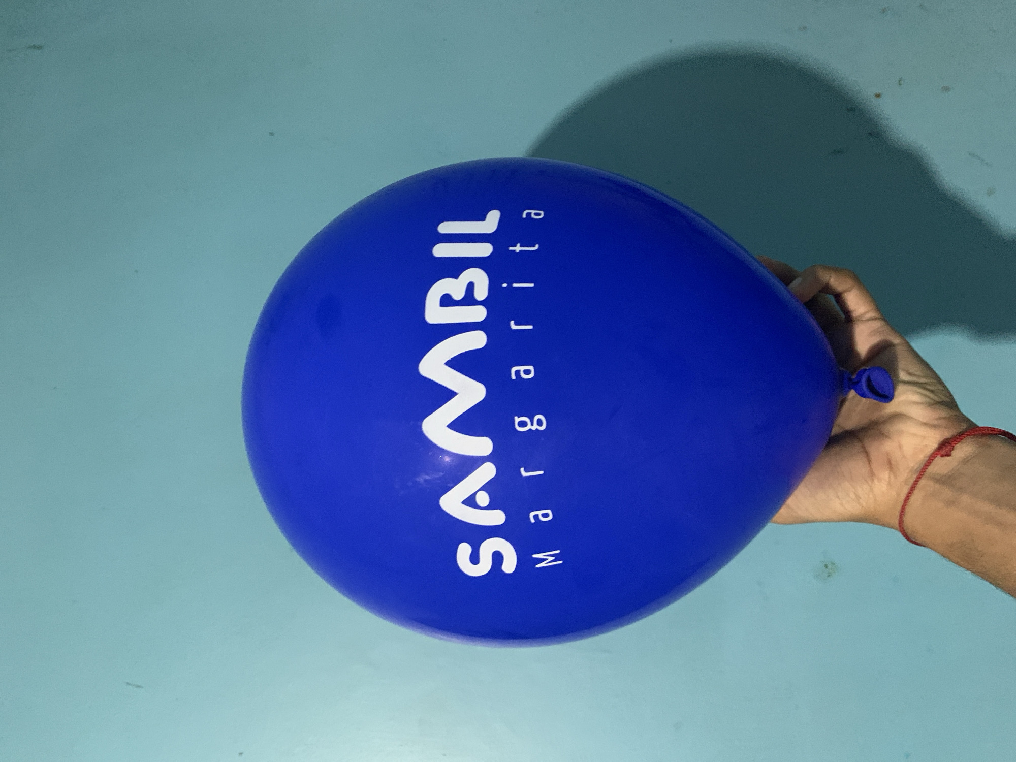 A blue balloon with the word "SAMBIL" in white letters stacked vertically above the word "Margarita" in smaller white letters is held in a person's right hand. The background is a light blue wall. The location is Santa Ana, Venezuela.