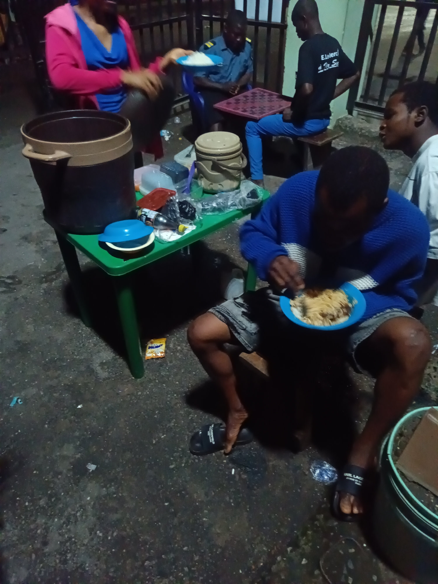 The image depicts a nighttime scene, likely in a public outdoor space in Ado, Nigeria, characterized by its uneven, worn asphalt ground. In the foreground, a person wearing a blue sweater and grey shorts is seated on a stool and eating from a blue plate containing noodles. To the left, a green plastic table holds a large dark brown water dispenser, a blue and white bowl, and other assorted items, including a red container, a bottle of liquid, and what appear to be packets of food. Behind the table, a woman in a pink jacket and blue top is interacting with the water dispenser. Further back, three men are gathered. One man, wearing a blue shirt, is seated at a small red and white checkered table, seemingly playing a game. Another man in a black t-shirt with white lettering is also seated. A third man, wearing a white t-shirt, is standing and looking towards the person eating. The overall impression is that of a casual gathering, possibly a meal or a game being played outdoors at night, with the environmental context suggesting a somewhat informal setting within a Nigerian city.