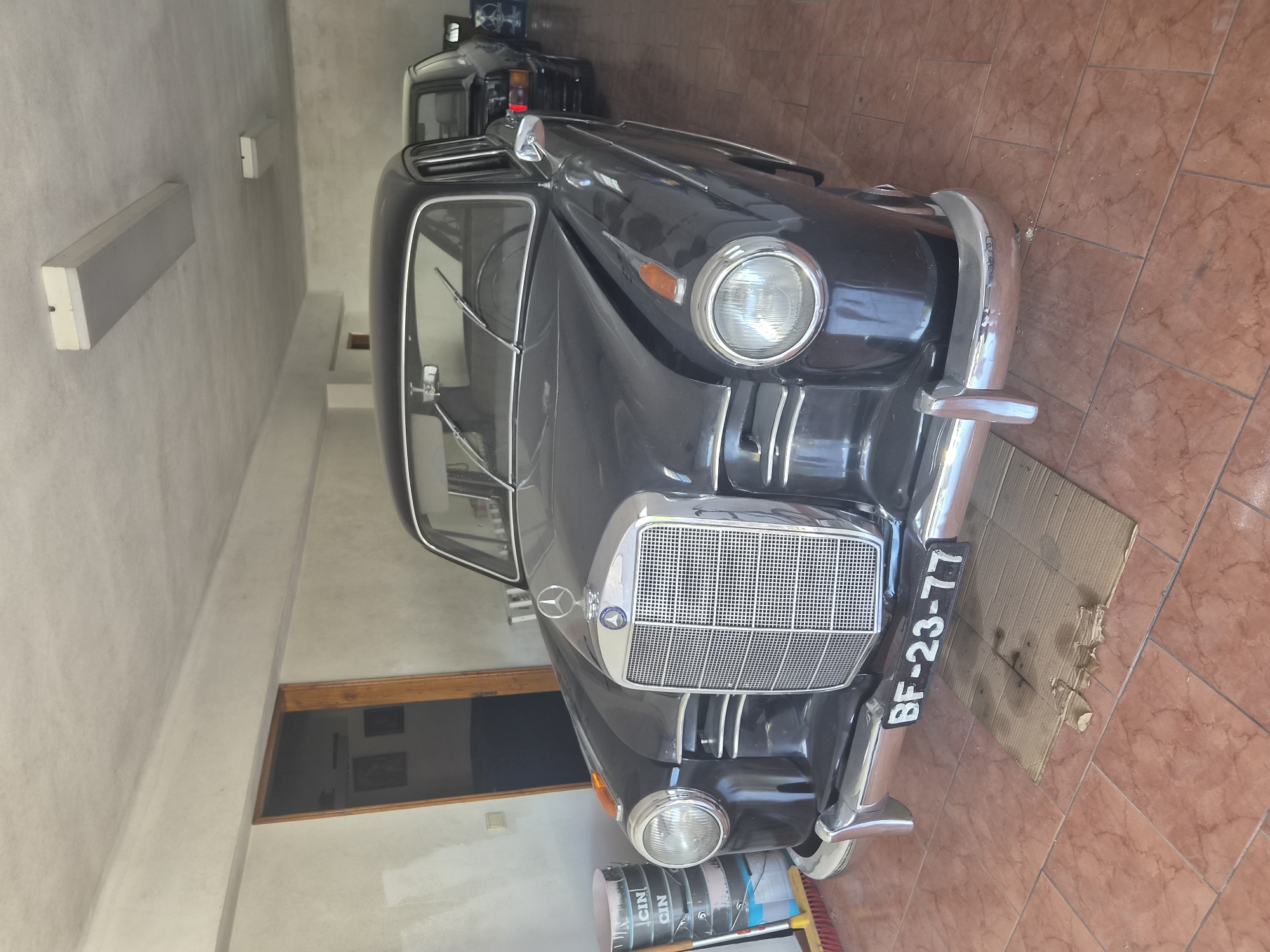 The image shows a vintage black Mercedes-Benz sedan parked indoors, likely in a garage or workshop in Seia, Portugal. The car is the primary subject, prominently displayed from the front. It features a chrome grille with the Mercedes-Benz logo on top, round headlights, and a license plate that reads "BF-23-77". To the left of the car, there are several stacked buckets, some bearing the label "CIN". A red-bristled broom leans against the wall behind the buckets. In the background, another dark-colored car is visible, and a door to an interior space is slightly ajar. The environment appears to be a tiled floor within a building that has fluorescent lighting fixtures on the ceiling. The overall scene suggests a storage or maintenance setting for classic automobiles.