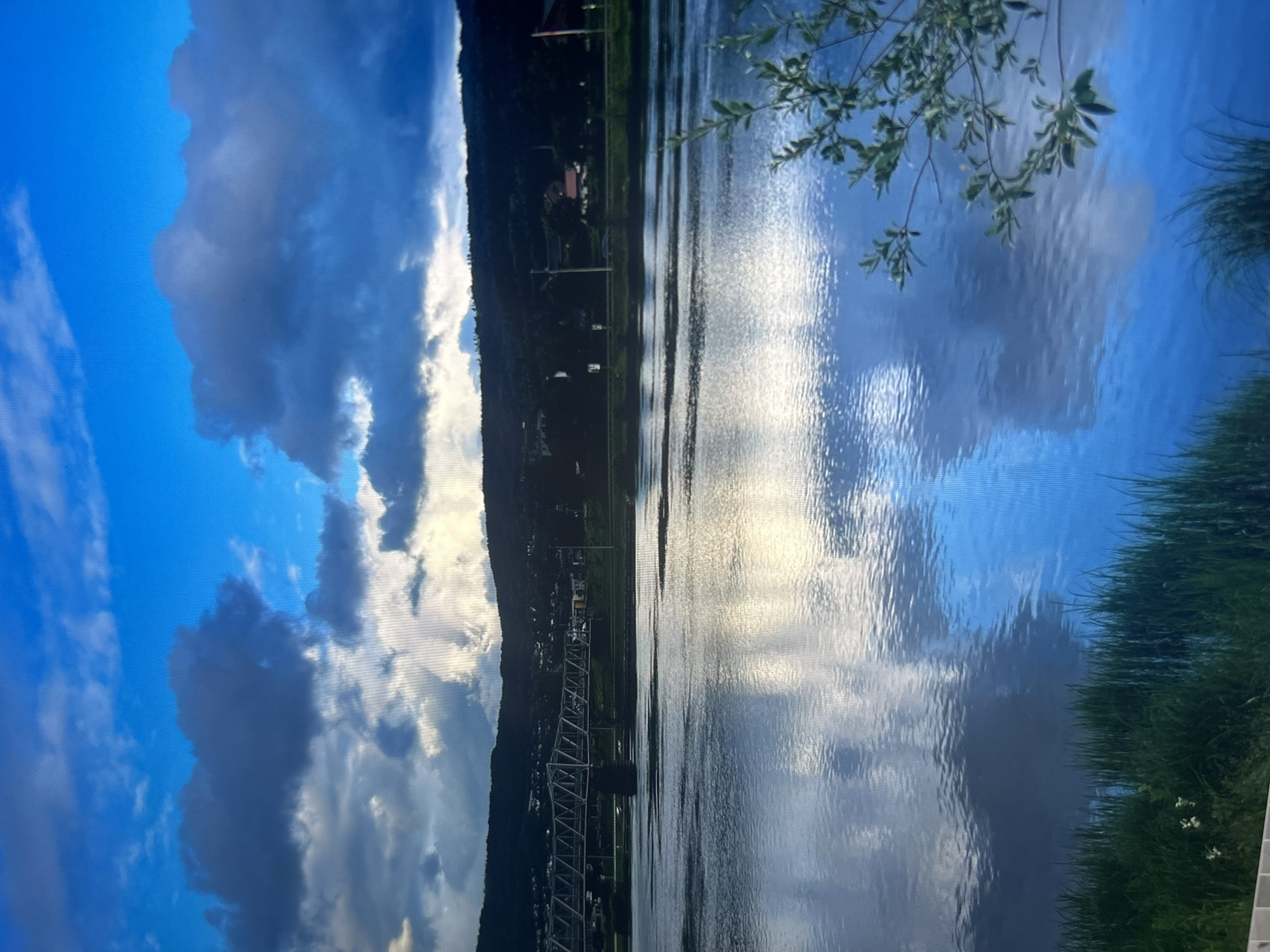 This is a photograph of a wide river with a reflective surface under a dramatic sky filled with dark clouds and patches of bright sunlight. In the distance, a long, metal truss bridge spans the river. Beyond the bridge and riverbank, a forested hillside rises, dotted with small buildings and houses. The foreground features lush green grass and a sprig of leafy branches on the right side of the frame. The overall impression is one of a scenic landscape with a bridge crossing a body of water, set against a backdrop of hills and a dynamic sky. There is no visible indication that this location is in Yagodovo, Bulgaria.