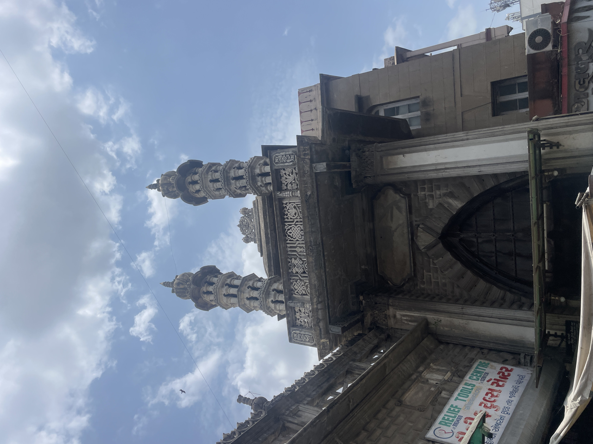 This low-angle, full-frame shot captures the ornate architecture of the Jama Masjid in Ahmedabad, India, under a partly cloudy sky. The main focus is the towering minarets, intricately carved with multiple levels of balconies and decorative finials. They flank a grand entrance with a large, arched doorway, surrounded by detailed stonework. The building appears to be constructed from a light-colored stone, with some areas showing signs of age and weathering.
To the left of the main structure, a sign for "RELIEF TOOLS CENTRE" is visible, indicating a commercial establishment integrated into or adjacent to the historical site. The sign is bilingual, with text in English and Gujarati. A bird is seen in flight in the upper left portion of the frame. The overall atmosphere is one of grandeur and historical significance, with a hint of modern commerce at the base of the ancient monument.