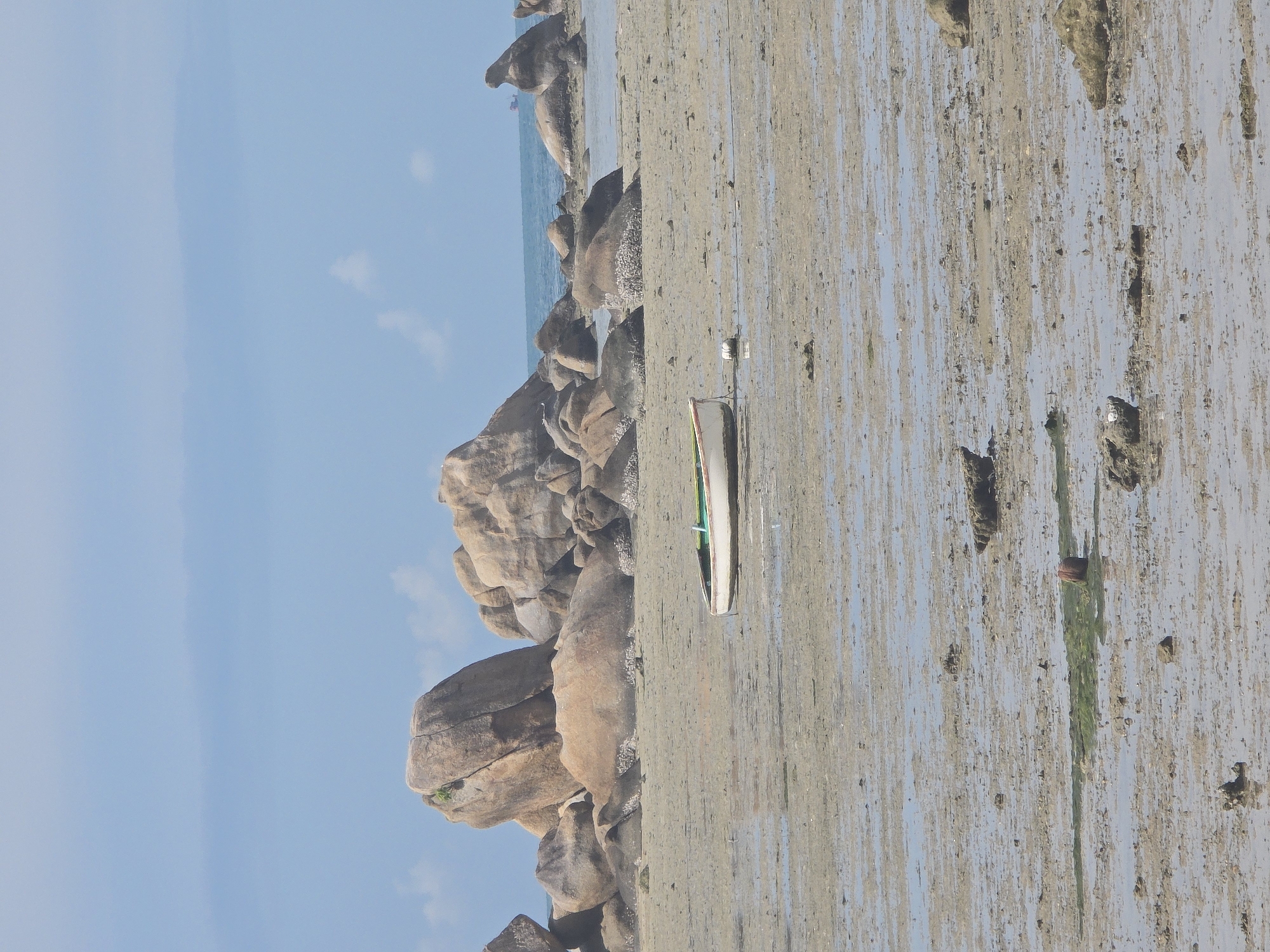 The image depicts a low-tide scene on a sandy beach in Baan Chaweng Noi, Thailand. A small, white boat with a green trim is resting on the wet sand near the water's edge. The beach is a mixture of sand and shallow water, with numerous rocks and boulders scattered across the landscape. In the background, larger rock formations are visible, with the ocean and a faint horizon line beyond them. The sky is a clear blue with a few wispy clouds. There are no people visible in the frame.