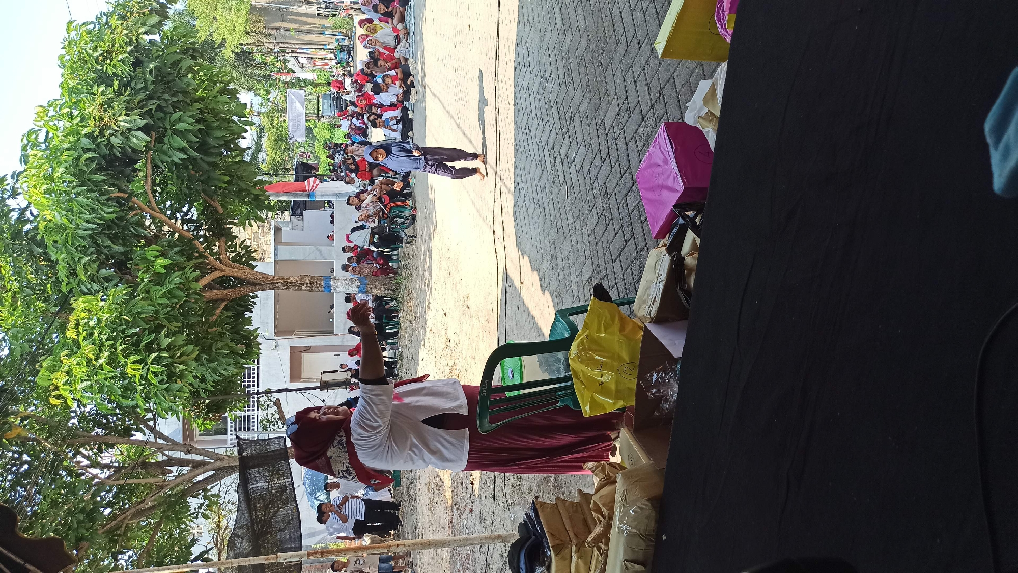 In the city of Galak, Indonesia, a group of people are gathered outdoors on a sunny day, possibly for an event or celebration. In the foreground, a woman wearing a white shirt and a maroon skirt and headscarf stands facing away from the camera, gesturing with her right hand towards a group of seated individuals in the background. A green plastic chair is positioned next to her. To her left, several cardboard boxes and bags are stacked.
In the middle ground, a young girl wearing a dark blue hijab and pants is walking. Behind her, a crowd of people, including many children, are sitting on the ground. They appear to be watching something or listening to someone. The background shows trees, buildings, and a street paved with interlocking bricks. The overall atmosphere suggests a community gathering.
