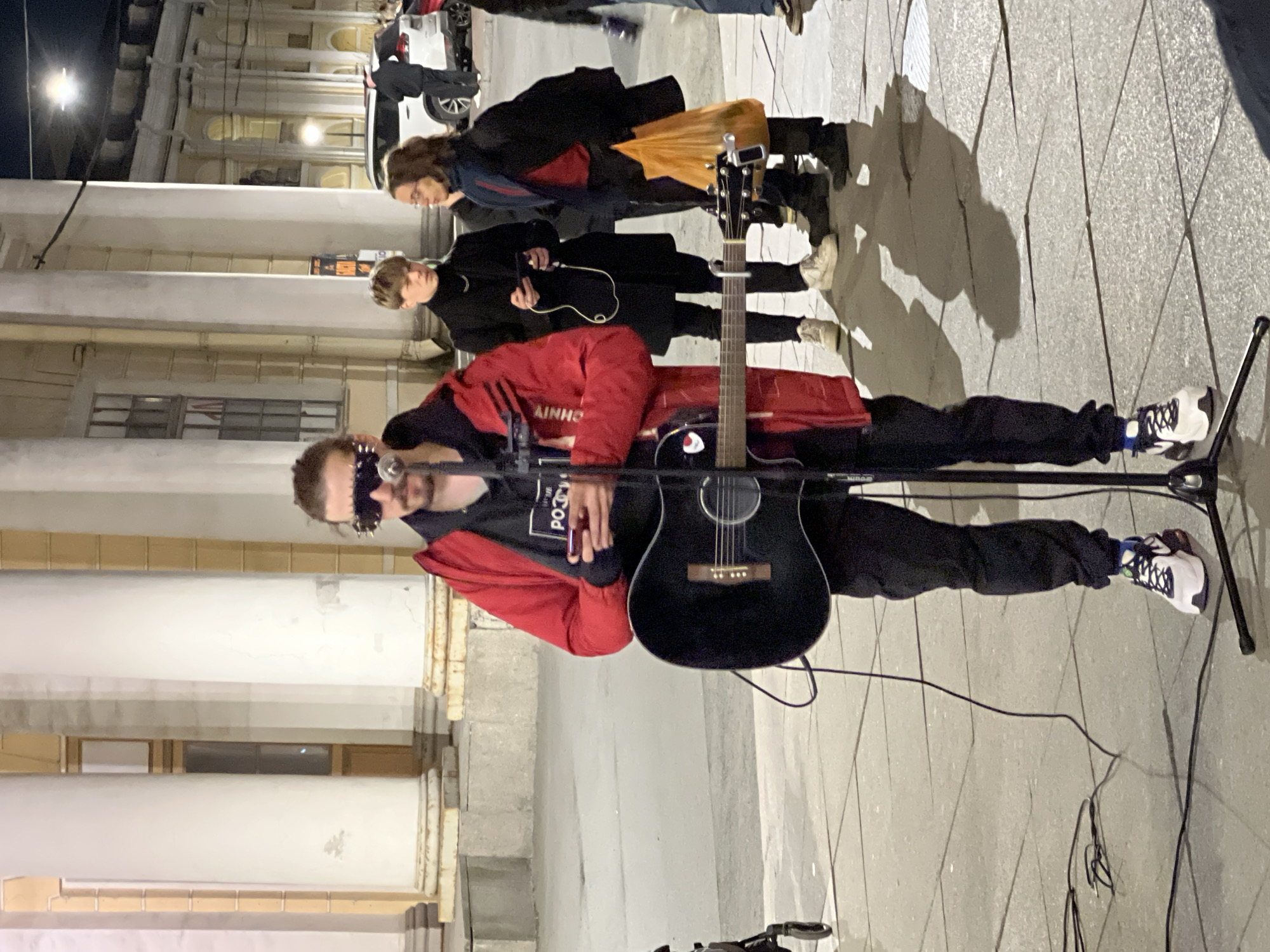 A street musician is performing in front of a building with columns, possibly in Saint Petersburg, Russia. The musician wears a red jacket, black t-shirt, black pants, and black and white sneakers. They have unique eye wear, appear to be standing with their guitar supported by a guitar stand, and holding a microphone. The guitar is black. Two people are observing the musician, with a third further away. Shadows are cast on the ground. A street light is visible in the background.