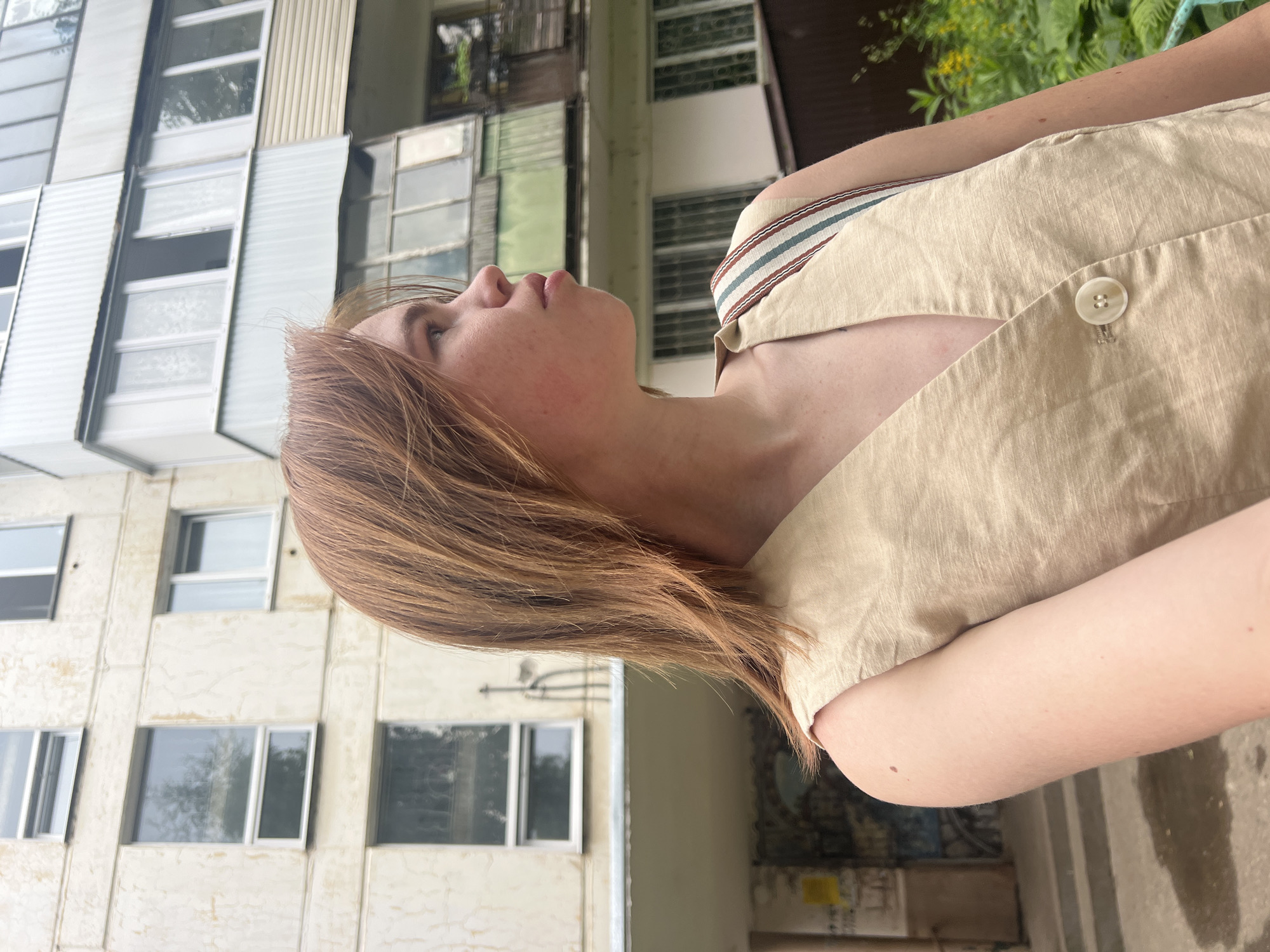 A young woman with reddish-blonde hair is standing in front of an apartment building, looking to her right. She is wearing a sleeveless beige vest with a V-neck and a single button. The building is a light beige color with multiple windows and balconies, some of which appear to be enclosed with glass. Greenery is visible to the right of the building. The overall setting suggests an urban environment, likely within the city of Chaikovsky, Russia, given the architectural style typical of Soviet-era apartment blocks.