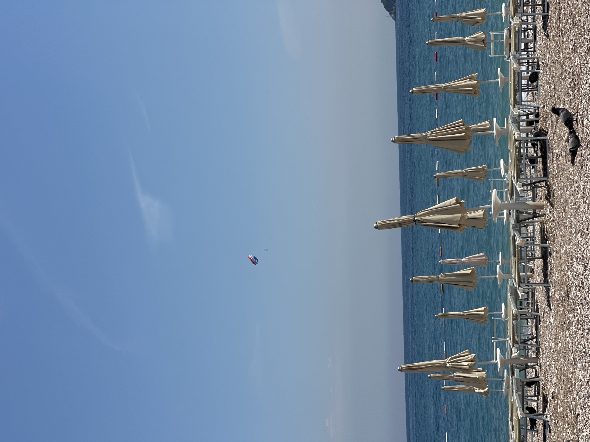 The image captures a vibrant and serene beach scene on a clear, sunny day, likely in Budva, Montenegro. The expansive sky, a brilliant blue with only a few faint wisps of clouds, dominates the upper portion. High in the sky, a colorful parasail, striped red, white, and blue, carries a single person, adding a dynamic element to the otherwise peaceful vista.
Below the sky, the calm, turquoise waters of the sea stretch towards a hazy horizon where a distant landmass is faintly visible. Scattered across the water are several small, red buoys, indicating swimming areas or boat boundaries.
Along the shore, a pebble beach is neatly lined with numerous rows of closed, beige beach umbrellas, each accompanied by white beach chairs and loungers. This organized arrangement suggests a resort or public beach setting, possibly captured during early morning or late afternoon before or after peak occupancy. Two dark pigeons are visible on the pebbly sand among the chairs, appearing to forage.
The bright, clear daylight indicates excellent weather, perfect for beach activities. The scene evokes a sense of calm and leisure, characteristic of a holiday destination. No visible text is present in the image.