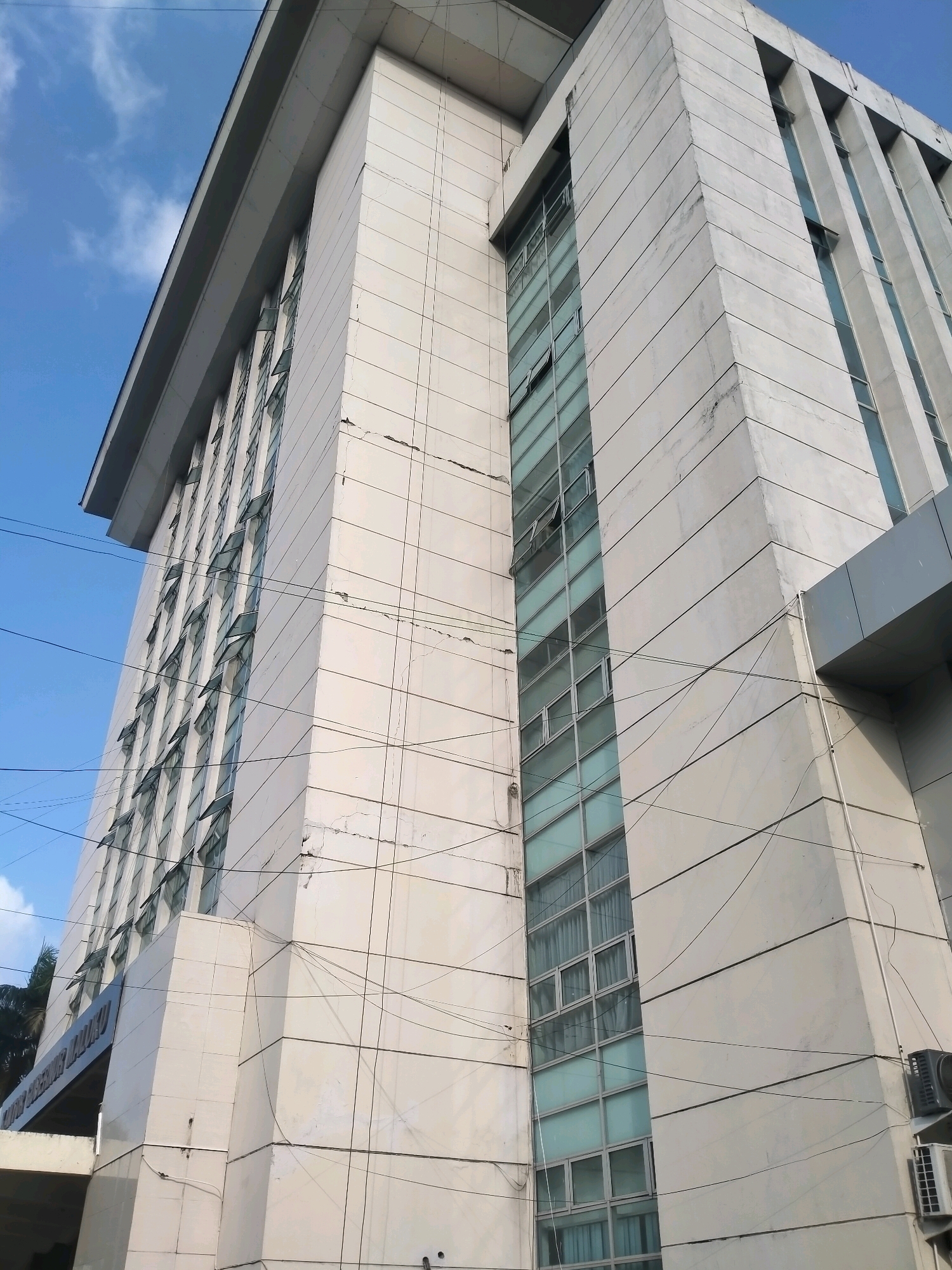 The image shows a low-angle view of a tall, modern building in Ambon, Indonesia. The building has a light-colored facade, likely made of marble or similar material, with horizontal lines and vertical columns. There are multiple rows of windows, some of which are open, with a tinted or frosted glass. The sky is blue with some scattered clouds. Overhead, several electrical wires crisscross in front of the building. On the right side of the building, an air conditioning unit is visible. The building appears to be an office building or a government institution, possibly the "Kantor Gubernur Maluku" (Maluku Governor's Office), as suggested by a partially visible sign at the bottom left of the image. The architecture is characteristic of urban development in Indonesia.
