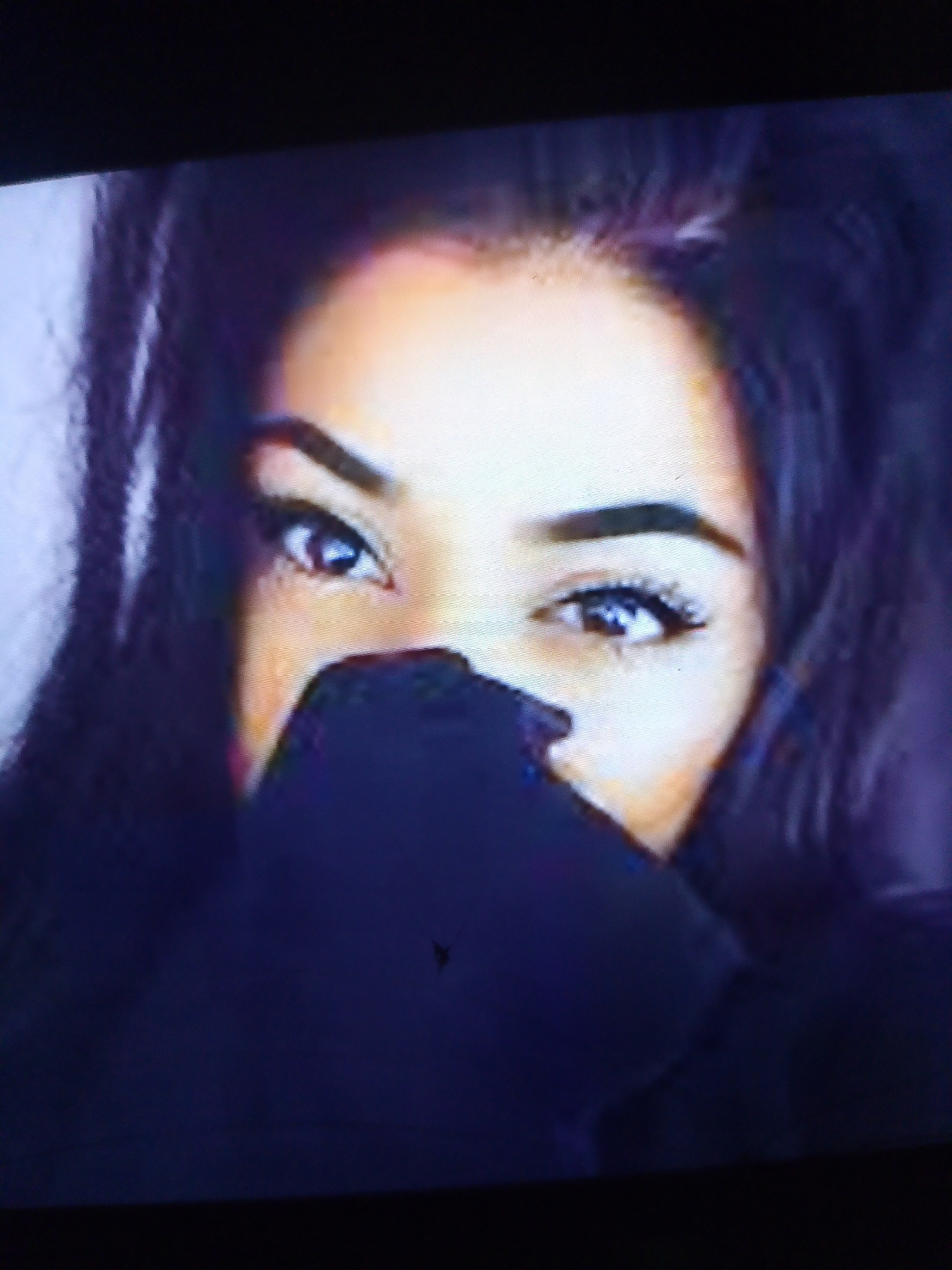The image shows a close-up of a young woman's face, with her mouth and nose covered by a dark blue garment, possibly a sweater or scarf. Her dark hair frames her face, and her eyes are visible, looking directly at the camera. Her eyebrows are well-defined, and her eyelashes are long and full. The lighting is soft, highlighting her skin tone. The background is a muted purple and blue. There is no discernible urban environment or specific location within Dobra Krynytsia, Ukraine, visible in the image. The image is a portrait of a person.