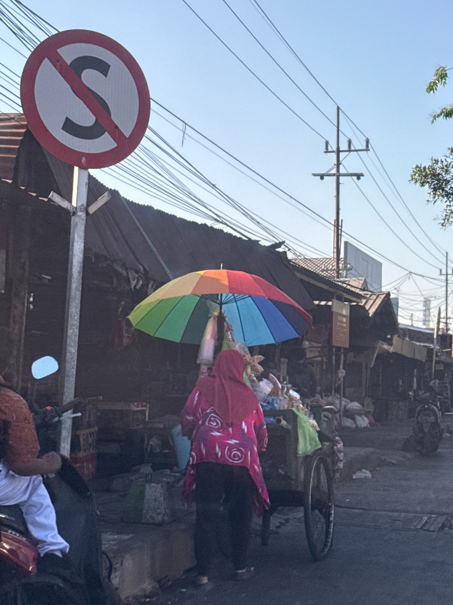 In Surabaya, Indonesia, a person wearing a pink headscarf and a patterned pink jacket pushes a cart adorned with a rainbow-colored umbrella and various goods. The cart is positioned on a street lined with shophouses that have corrugated metal roofs. A "No Stopping" sign is visible on the left side of the frame, indicating traffic regulations. Power lines crisscross the sky, and a utility pole stands in the background, common sights in urban Indonesian landscapes. The scene suggests a bustling street market or roadside commerce.