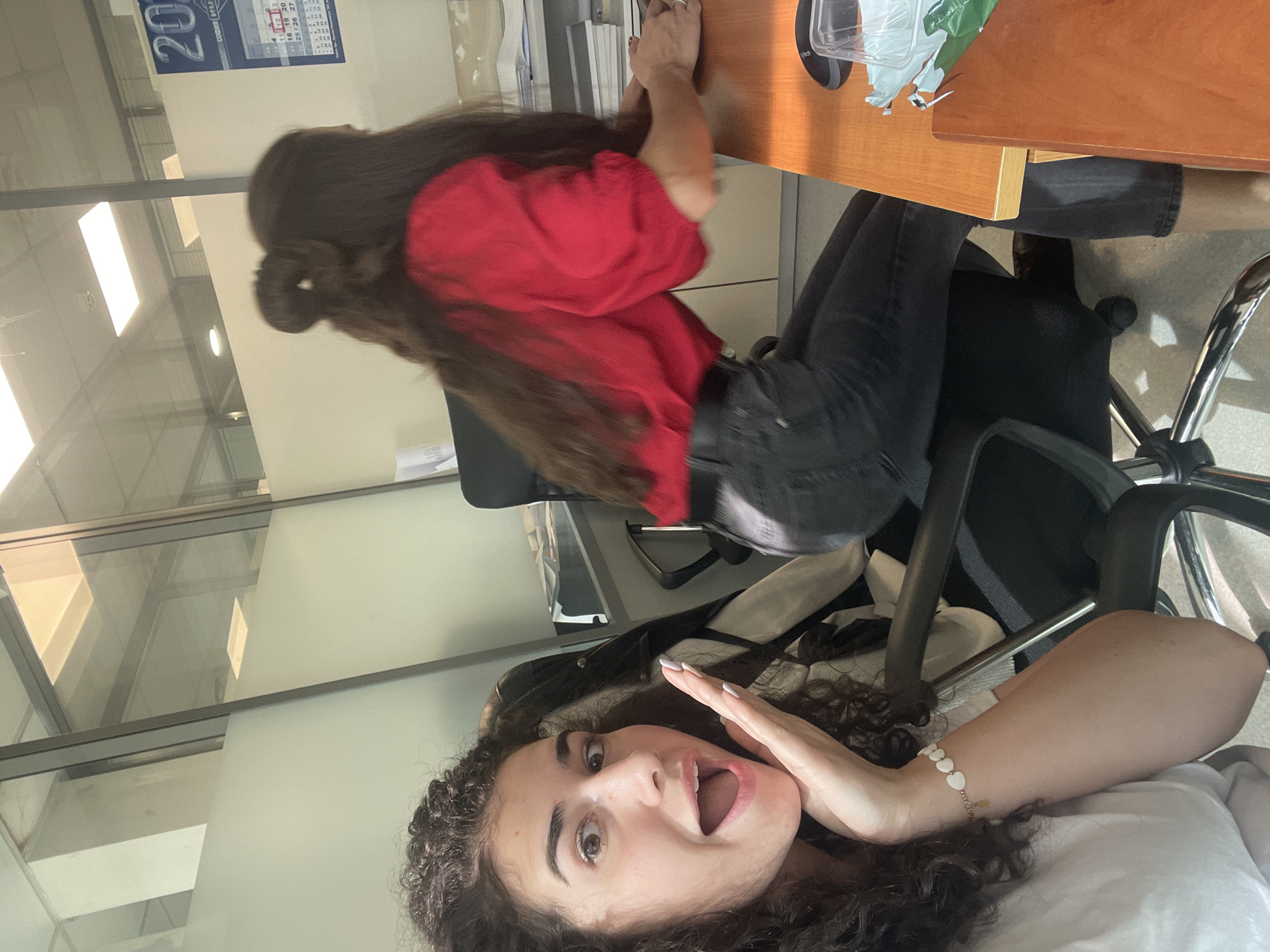 Two women are in an office. One is seated at a desk and facing it with a red shirt and black pants. The second woman is looking directly at the camera, with a surprised expression, in a white shirt. The office has large windows with a view. A calendar is visible on the wall. This is taking place in an office, most likely in Sofia, Bulgaria.