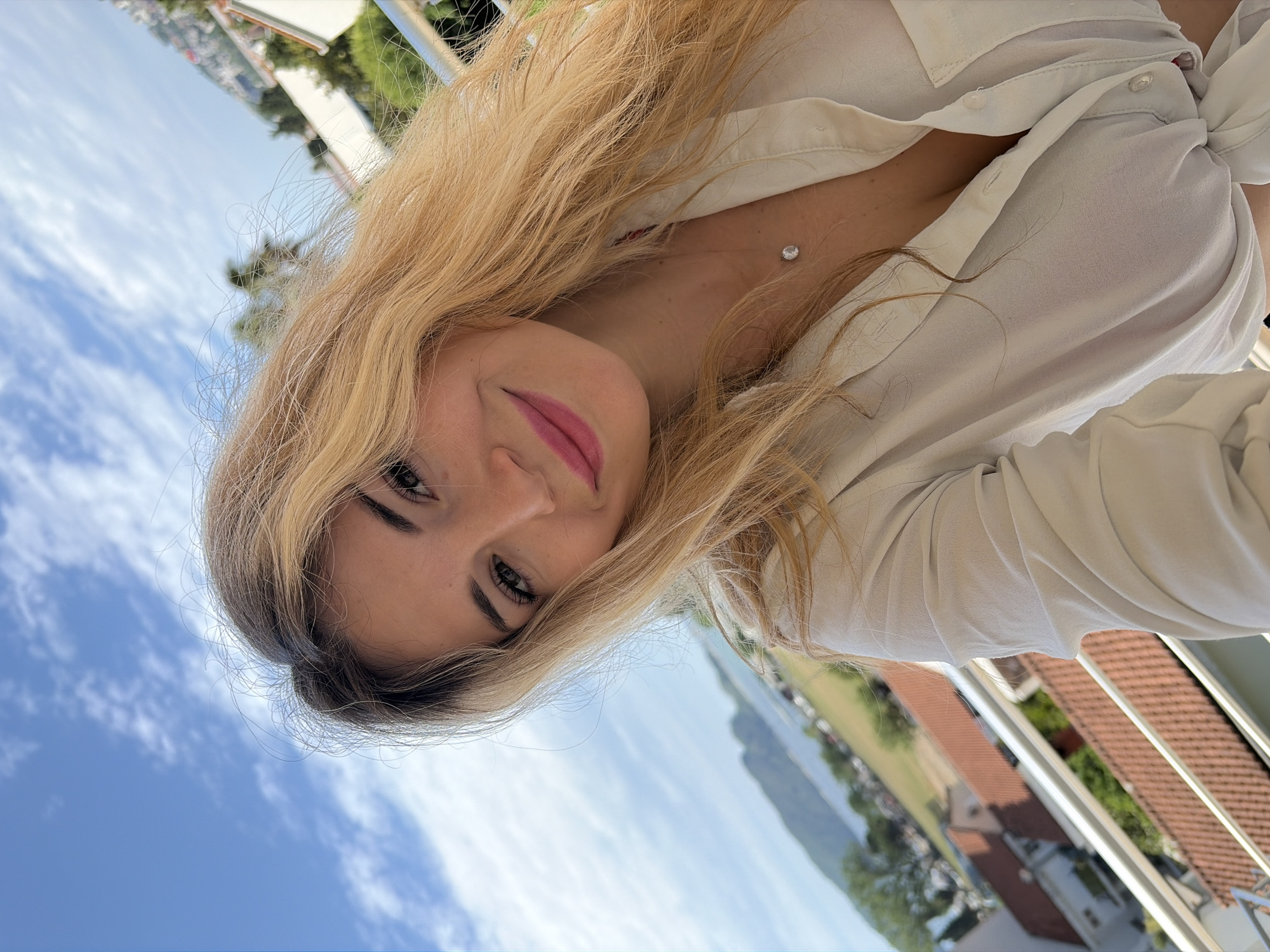The image is a selfie of a woman with blonde hair wearing a white shirt, taken outdoors on a balcony. Her head is tilted, and she is looking directly at the camera with a slight smile. The background shows a cityscape, likely in a mountainous region. The blue sky with some clouds is also visible. Based on the architecture and landscape features, it's plausible that the location is Budva, Montenegro, a coastal city known for its mountains and beaches. The balcony has a metal railing.