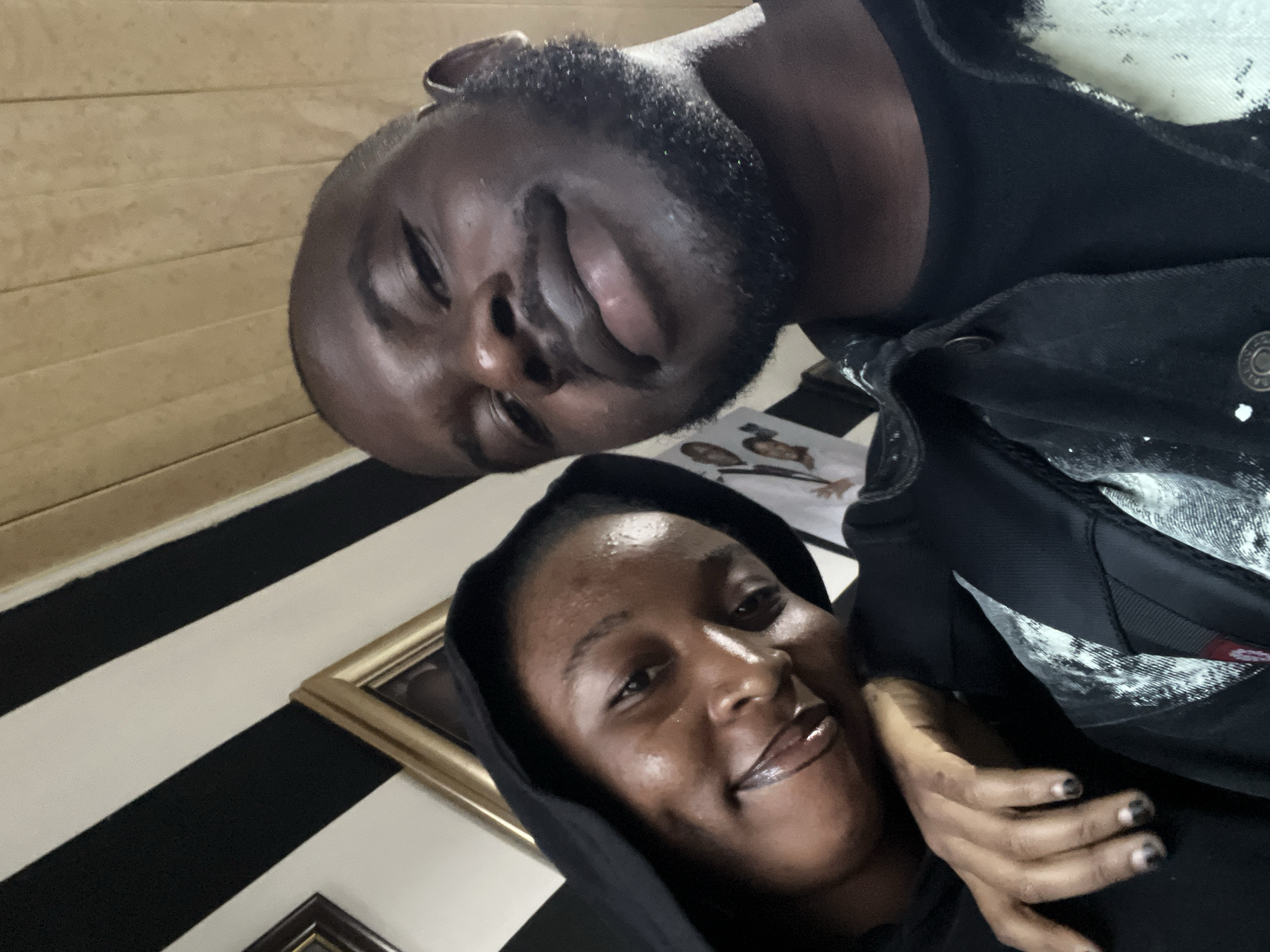 This image depicts two dark-skinned individuals, a female on the left and a male on the right, captured in a close-up indoor selfie in Jos, Nigeria.
The female subject, positioned on the left, wears a dark hooded garment. Her head is tilted slightly, and she displays a soft, closed-mouth smile. Her lips appear glossy with a dark liner, and her skin shows a subtle sheen. Her visible right hand rests near her chest, revealing fingernails painted with a dark color.
The male subject, occupying the right side of the frame, has a closely cropped haircut, a mustache, and a short, well-maintained beard. He wears a plain dark t-shirt beneath a dark denim jacket that features white paint splatters or a distressed pattern. Metal buttons are visible on the jacket's front. He holds the camera, looking directly into the lens with a slight, closed-mouth smile.
The female is leaning her head gently against the male subject's left shoulder, indicating a relaxed interaction.
The background elements suggest an indoor, decorated space. To the upper left, diagonal black and white stripes are visible on a wall. Below this, a portion of a dark wooden picture frame is present. Above the female's head, part of a golden picture frame is visible. To the upper right, above the male's head, a section of light brown wooden paneling with vertical slats can be seen. Behind the male, a white surface with faint images of faces is partially visible.