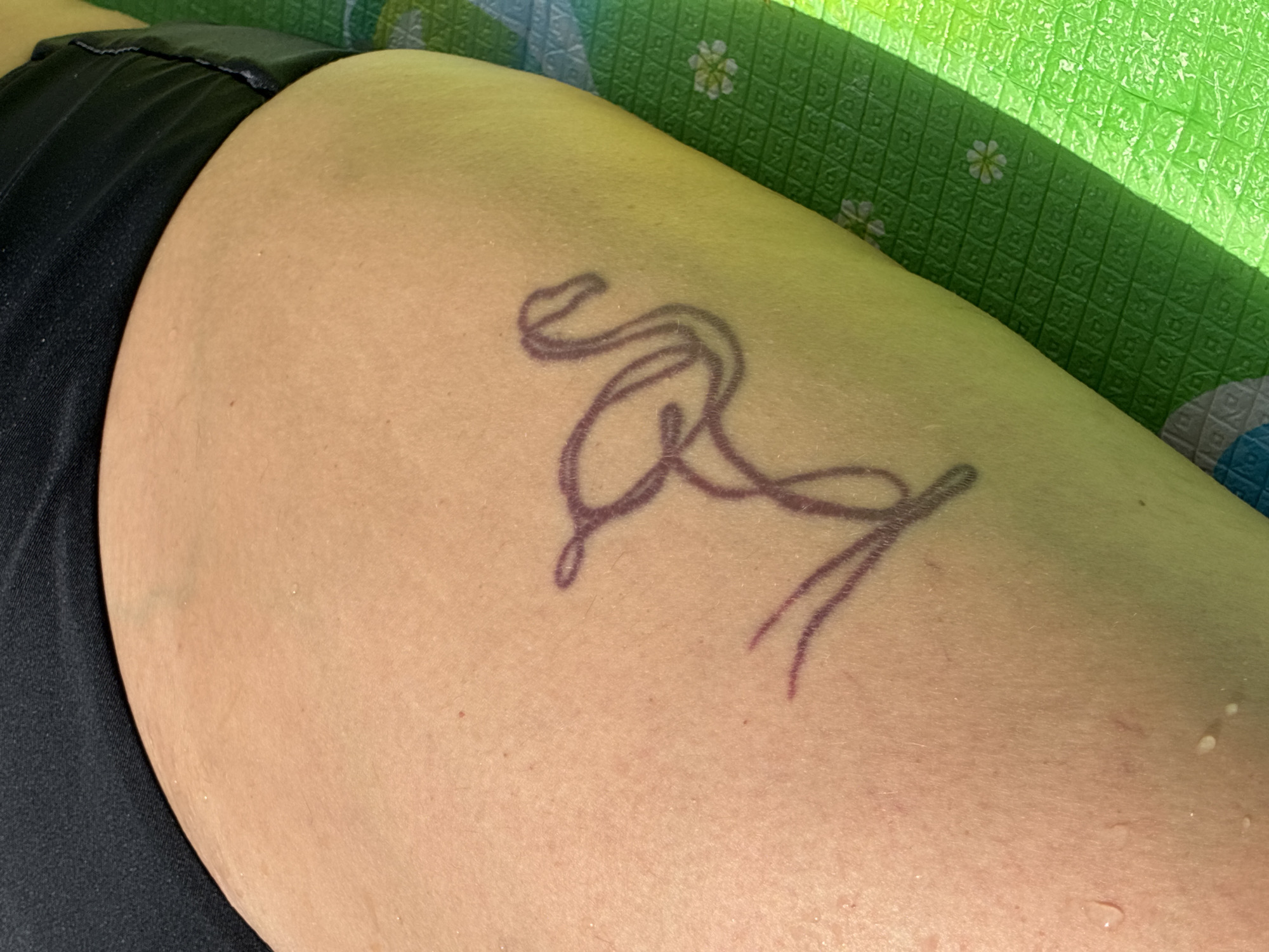 A close-up, medium shot captures a portion of a person's inner thigh, likely a woman's given the black fabric of swimwear at the top left of the frame. The skin, appearing tan, is slightly damp with small water droplets visible. A stylized, outline tattoo of a flamingo in a purplish-brown ink is inked on the thigh. The flamingo is depicted in a flowing, minimalist style. The skin is resting on a green mat with a textured grid pattern and small white flower motifs. The lighting suggests an outdoor setting, possibly in a warm climate. The location in Surami, Georgia is not explicitly depicted in the image.