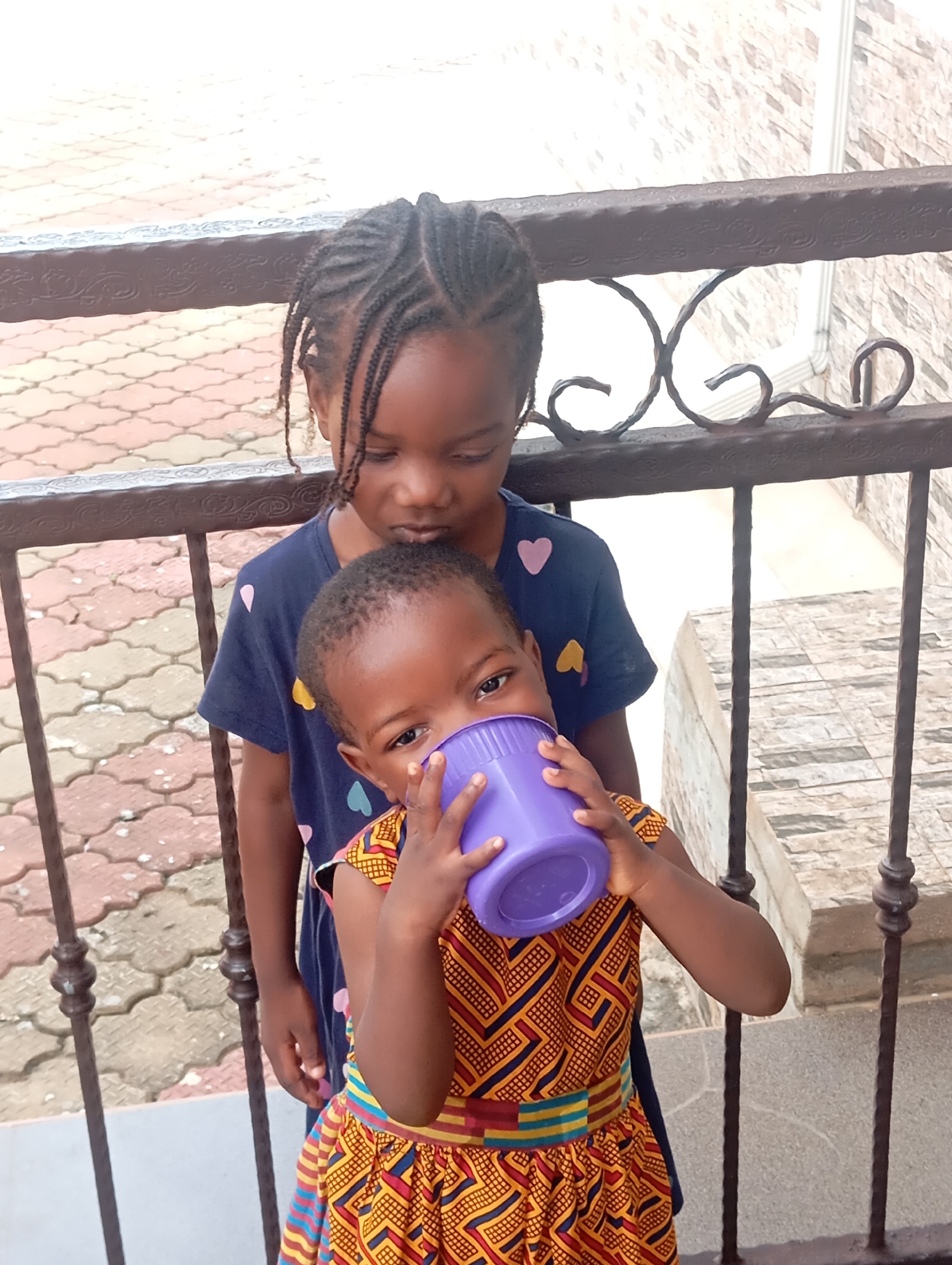 The image captures two young Black girls standing behind a dark metal railing. The girl in the foreground, wearing a vibrant yellow and red patterned dress with a colorful striped belt, is holding a purple plastic cup to her mouth, seemingly drinking from it. Her eyes are visible and looking towards the camera. Behind her, the older girl, dressed in a dark blue t-shirt adorned with small heart-shaped prints, looks down towards the younger girl. Her hair is styled in numerous braids. The railing is ornately decorated with curved metalwork. In the background, paving stones with a patterned design are visible, along with a stone-tiled wall and a white downspout. The scene appears to be outdoors, possibly on a balcony or patio. The location is identified as Nsugbe, Nigeria.