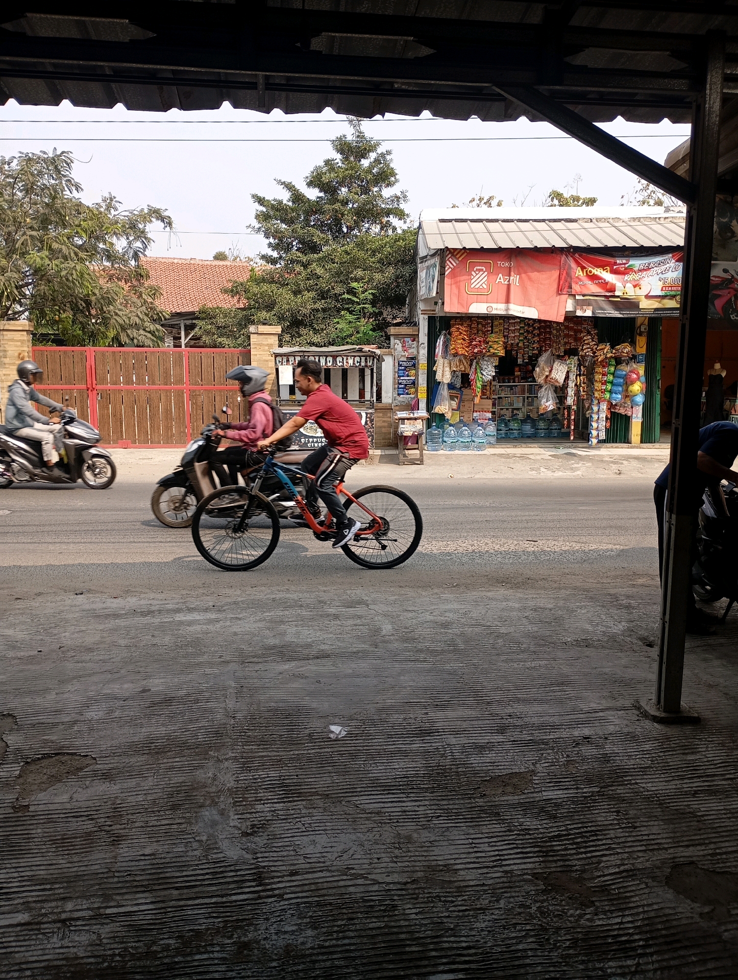 The image depicts a street scene in Babelan, Indonesia, with a focus on a cyclist and two motorcyclists. In the foreground, a man in a red shirt is riding a bicycle. He is positioned on the right side of the road, moving towards the left of the frame. He is wearing a dark helmet and dark pants. Behind him, a motorcyclist wearing a pink shirt and a dark helmet is also riding a motorcycle. Further to the left, another motorcyclist in a grey jacket and helmet is riding a scooter. The road surface appears to be asphalt, with some wear and tear.
To the right of the road, there is a shop with merchandise displayed outside, including bags of snacks and bottled water. The shop has a red awning and a sign with Indonesian text. A portion of a building structure with a dark roof and support beams is visible in the immediate foreground on the right, obscuring part of the shop.
In the background, there are trees and residential buildings with red tile roofs. The sky is clear and bright, suggesting it is daytime. The overall impression is of a typical commercial and residential street in a local Indonesian community.