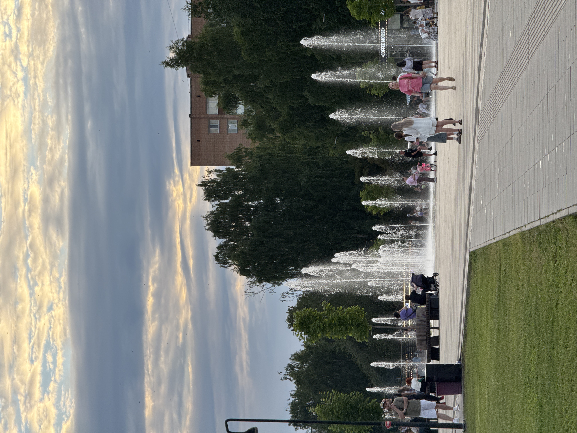 The image depicts a public square with a large fountain in the background, illuminated by spray. Several people are visible, some walking and others sitting on benches. A stroller is present. The area is paved, with a grassy area to the left. Trees and a brick building are seen behind the fountain. The sky is cloudy with patches of sunlight. The location is identified as the Central Square of Izhevsk, Russia.