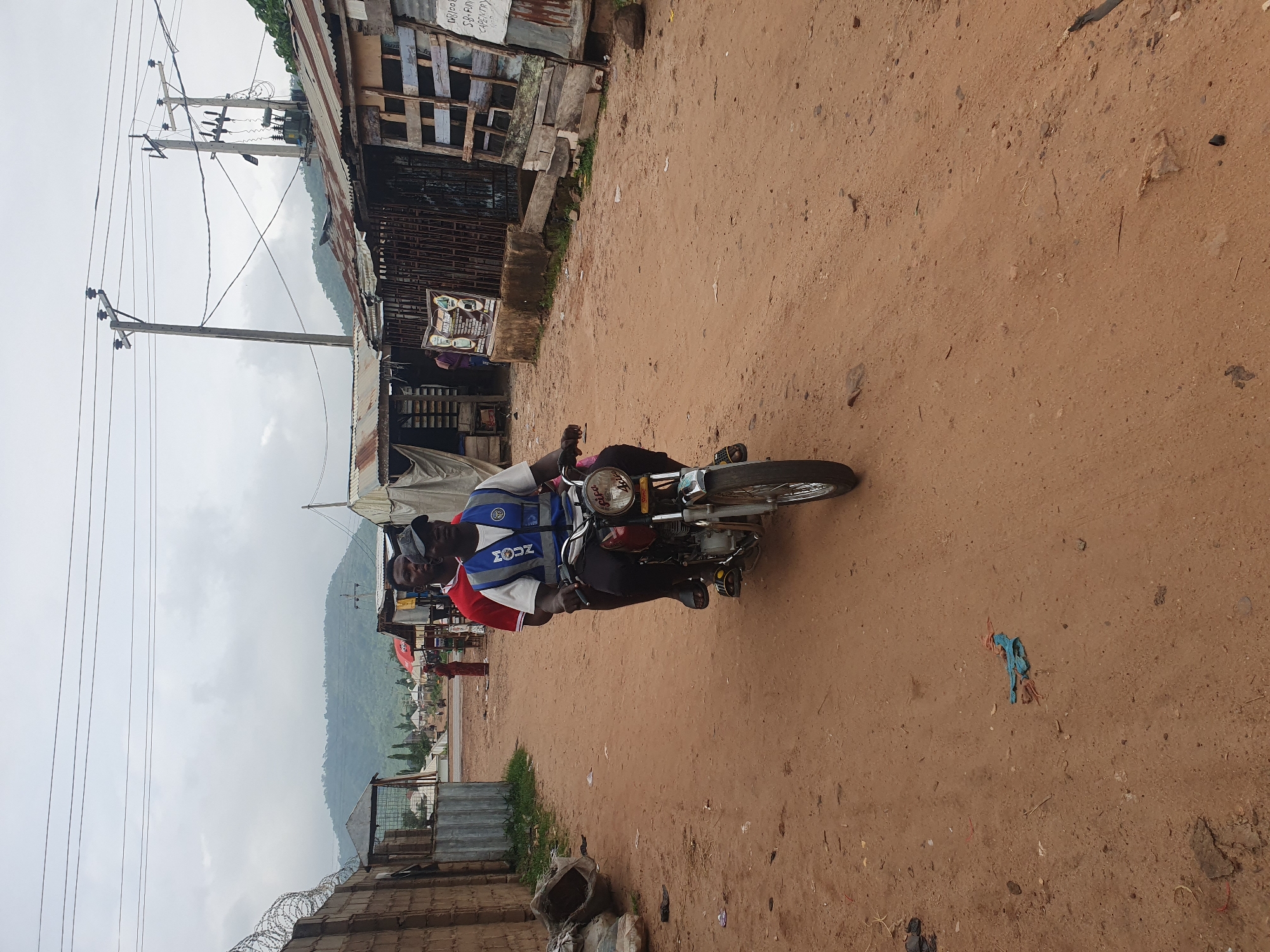 The image depicts a street scene in Lokoja, Nigeria, under an overcast sky. In the foreground, two men are riding a motorcycle on a dusty, reddish-brown dirt road. The man in front is wearing a blue vest with white lettering that reads "MOUNT." He is also wearing a white t-shirt and dark pants, and a dark cap. The man behind him is wearing a red shirt. The motorcycle is dark red and appears to be older model.
To the left of the road, a textured brick wall topped with barbed wire runs along the edge of the scene, with some overgrown vegetation and discarded sacks. Further down the road on the left, a gate-like structure with a metal roof is visible.
To the right of the road, a series of ramshackle structures with corrugated iron roofs line the street. These buildings appear to be shops or market stalls, with some displaying goods or signage. One sign is partially visible and seems to advertise services like carpentry. A transformer and utility poles with electrical wires are present above these structures.
In the background, the road continues, leading towards a line of buildings and further into a more distant landscape characterized by green hills and trees. Other people are visible in the distance, some standing and others possibly walking. The overall impression is that of a bustling but somewhat unkempt urban or peri-urban environment in Nigeria.