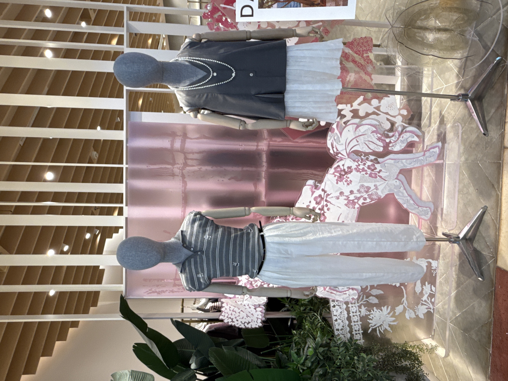 The image shows two mannequins in a store display. The mannequin on the left is wearing a striped sleeveless polo shirt and white wide-leg pants, accessorized with a black belt. The mannequin on the right is wearing a dark grey sleeveless vest over a white pleated skirt, and a long pearl necklace. Both mannequins are grey and headless. They are displayed in front of a pink, shimmering backdrop with a decorative pattern of stylized flowers and animals. To the left of the display is a large green plant. The floor is tiled with a geometric pattern. A sign with the letter "DA" is visible to the right of the display. The overall setting appears to be an indoor retail space, possibly a fashion boutique. There is no specific indication of the city of Huizhou, China.
