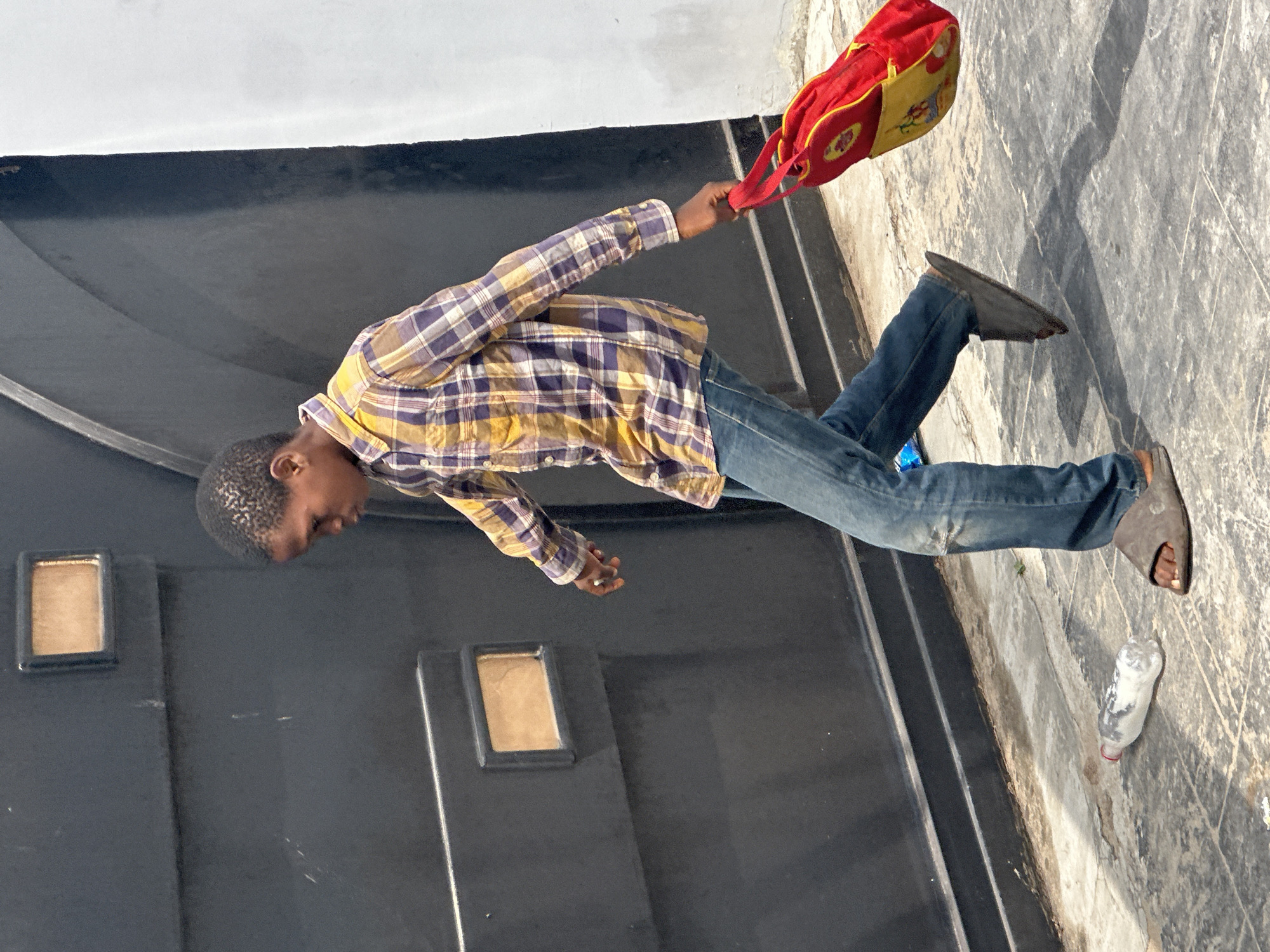 A young Black boy, appearing to be of school age, walks on a paved walkway next to a large, dark industrial-style metal gate. He is wearing a plaid shirt in shades of yellow, purple, and white, blue jeans with a slight tear on the left knee, and dark gray open-toed sandals. In his right hand, he holds a red and yellow bag with a cartoonish graphic. To his left, on the ground, is a discarded plastic water bottle with a red cap. The environment suggests an urban setting, possibly in Badagry, Nigeria, given the architecture and the boy's appearance. The lighting indicates it is daytime, with distinct shadows cast by the boy and the bag.