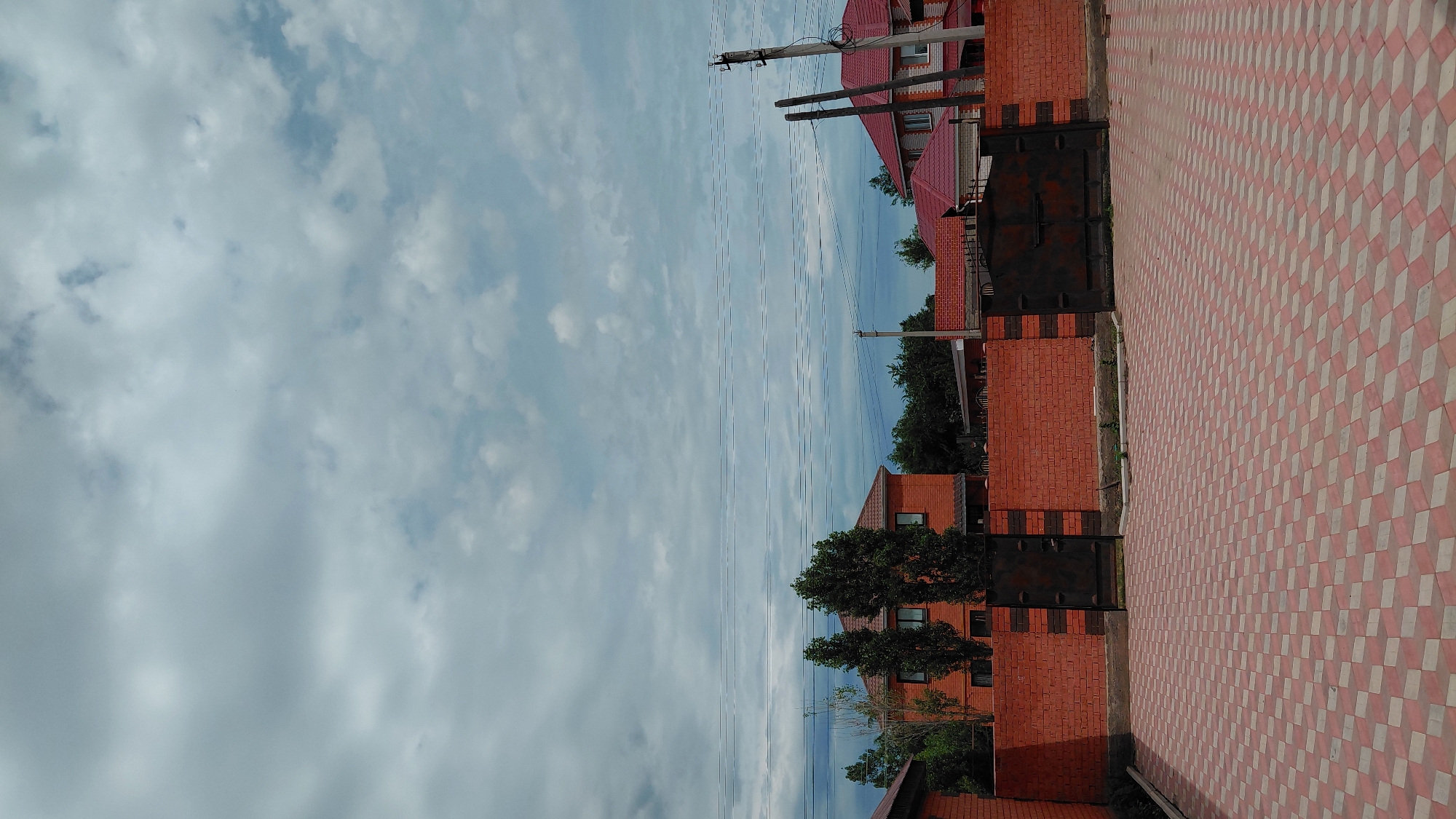 The image depicts a residential area under a cloudy sky, likely in Aktobe, Kazakhstan. In the foreground, a paved courtyard with a diamond pattern of red and grey tiles is visible. A low, red brick wall with dark metal gates stretches across the middle ground, dividing the courtyard from the properties behind it. Several mature trees stand behind the wall, their green foliage contrasting with the brickwork. In the background, two distinct houses are partially visible. The house on the left is a two-story structure with a reddish-brown brick facade and dark windows. To its right, another house features a red tiled roof and a lighter colored upper story with white trim. Utility poles and overhead wires are also present in the scene. The overall impression is of a suburban or semi-rural environment.