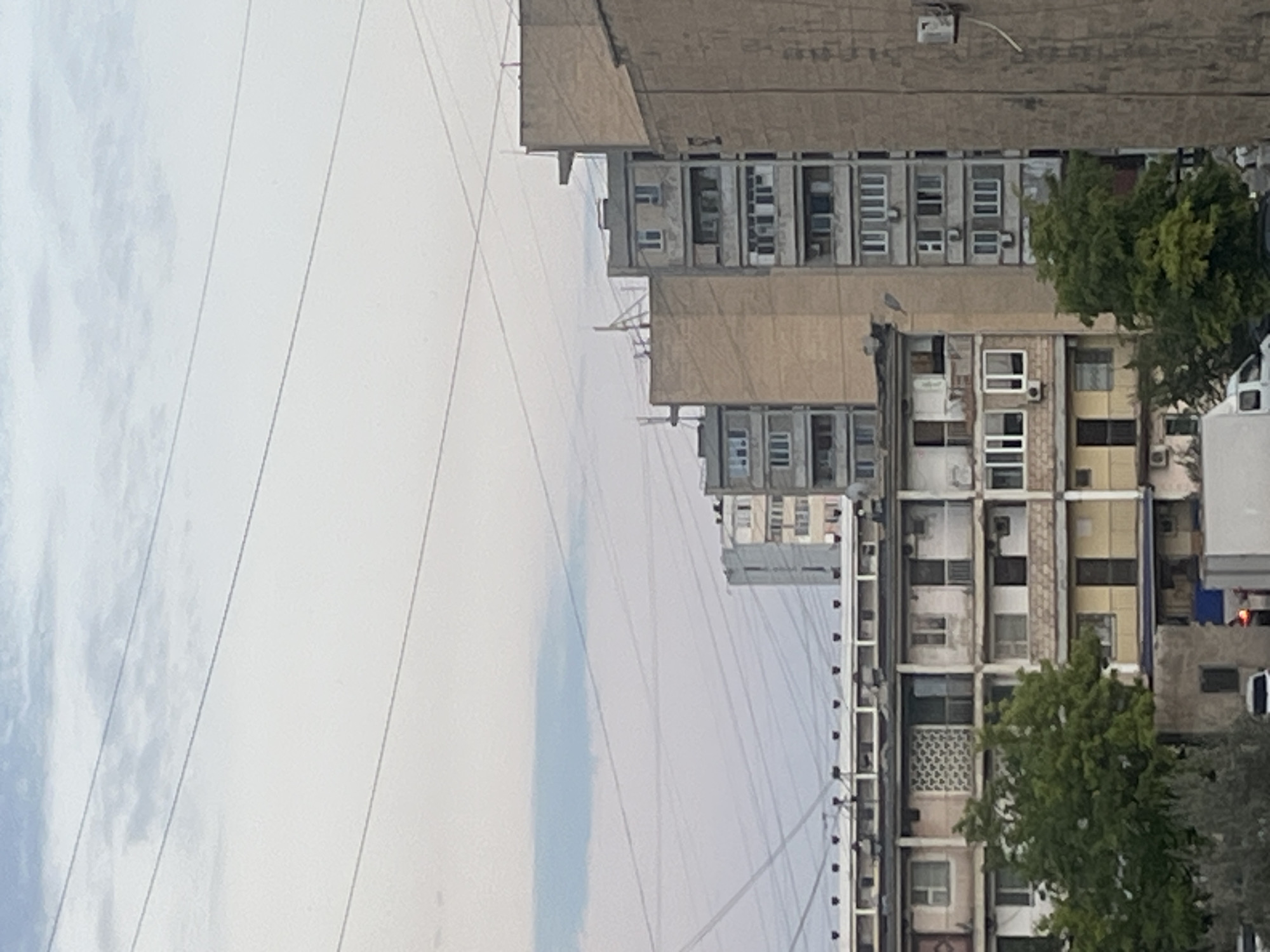 This media file captures a section of a city, oriented vertically and viewed from a slightly elevated perspective. The scene is dominated by several multi-story buildings, characterized by their utilitarian architectural style. These structures feature numerous windows, some with air conditioning units visible, and a mix of concrete and lighter-colored facade materials. Power lines stretch across the frame, creating a diagonal pattern against the sky. A large, leafy green tree is prominent in the middle-ground, partially obscuring one of the buildings. The sky appears overcast or hazy, suggesting diffused daylight. There are no people visible in the image. The setting is urban, with a density of buildings typical of a residential or mixed-use area. The overall impression is one of a functional, perhaps older, urban environment.