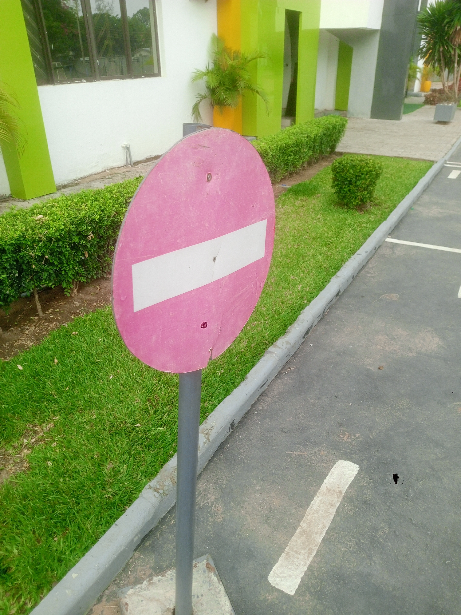 The image depicts a pink "do not enter" sign posted beside a gray paved area in what appears to be a landscaped parking lot or driveway. The sign is positioned between the paved area and a strip of green grass bordered by bushes and a curb. The background features a building with sections painted in green, yellow, white, and gray. A potted plant is visible near the building. The location is likely in Abuja, Nigeria, based on the architectural style and vegetation, but there is no definitive identifier in the frame. There are no people or animals visible in the image.