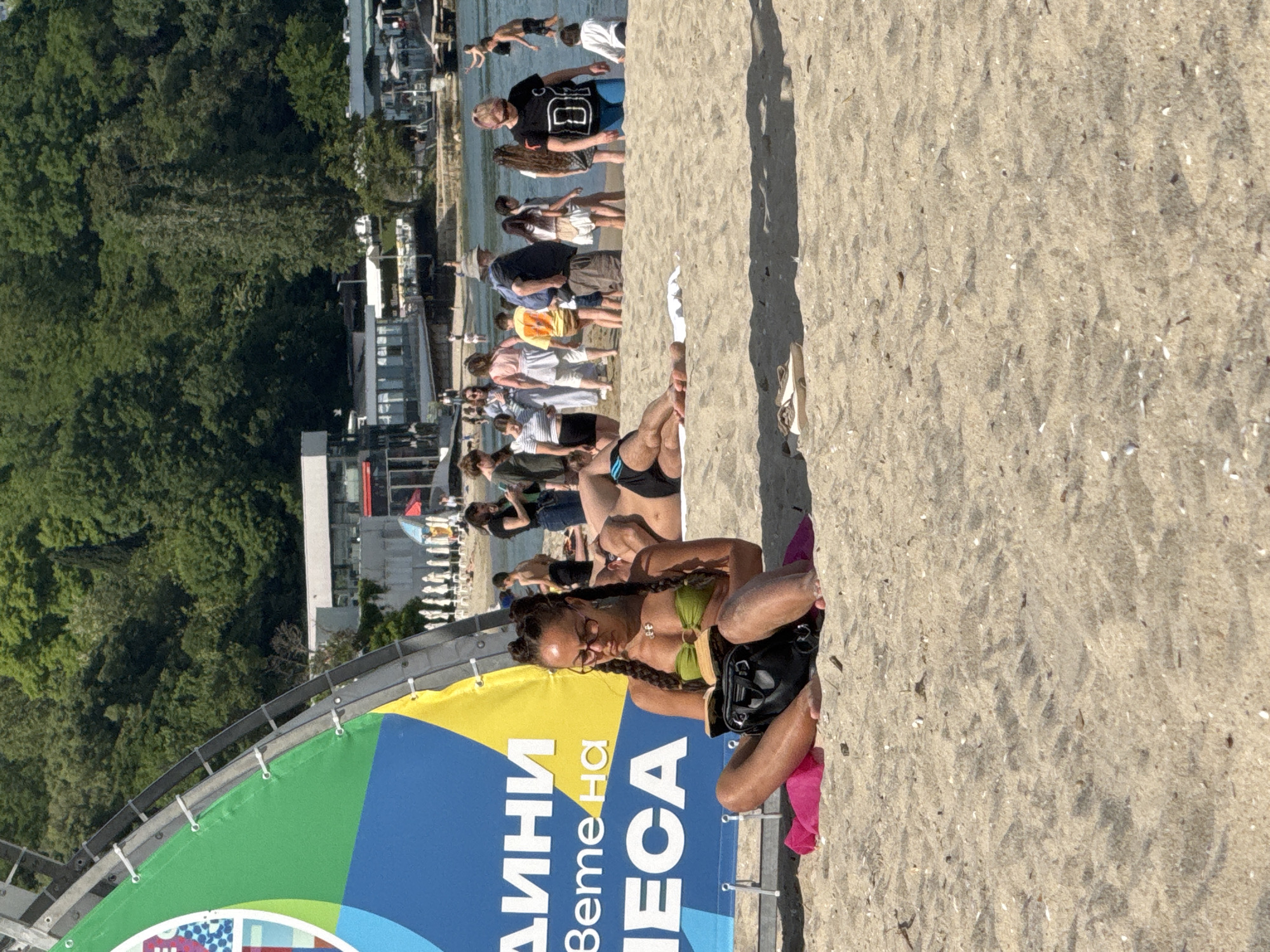 Here is a detailed description of the image:
**Scene:** A beach in Varna, Bulgaria. The focus is on a woman sitting on the sand near a large banner.
**People:**
* A woman with braided hair, wearing glasses and a green bikini top, is sitting cross-legged on the sand. She is holding a black bag or purse.
* Several other people are visible in the background, walking or standing along the shoreline. Some are swimming in the water.
**Objects:**
* A large banner with text in Bulgarian and a mix of green, blue, and yellow colors.
* A black bag is held by the woman in the foreground.
* Sand and some small debris or shells scattered on the beach.
**Actions:**
* The woman is sitting on the sand, possibly relaxing or taking a break.
* People in the background are swimming, walking, or standing near the water.
**Interactions:**
* The primary subject is the woman sitting near the banner. There appear to be some interactions between the people walking in the background.
**Environment:**
* The scene is an outdoor beach environment.
* In the background, there is a treeline and buildings visible near the shore.
* The sky is not visible, but the lighting suggests it is daytime.