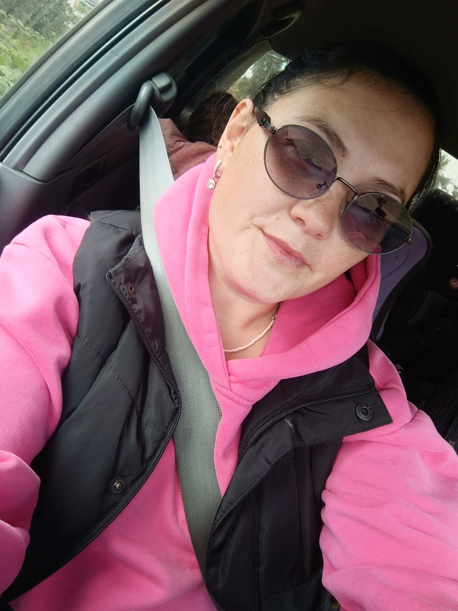 A medium shot captures a woman sitting in the driver's seat of a car, with a seatbelt fastened across her chest. She is wearing round, tinted sunglasses, a pink hooded sweatshirt, and a black vest. Her hair is dark and pulled back. The interior of the car is visible behind her. The lighting suggests it is daytime. There is no specific indication of the city of Karsha, Russia, in the image.