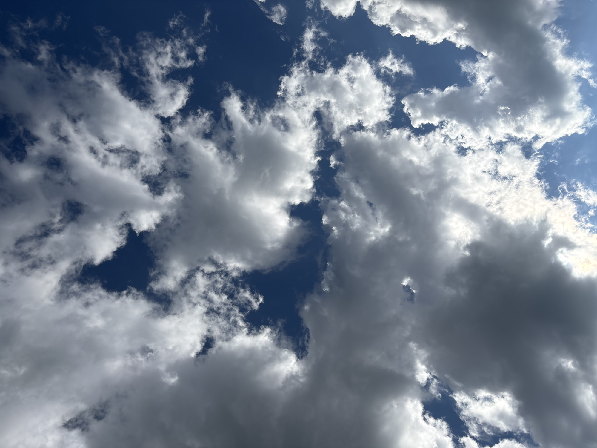 A high-angle shot captures a dramatic sky filled with a mix of dark, heavy clouds and bright, fluffy white clouds against a deep blue sky. Sunlight is breaking through in several areas, illuminating the edges of the white clouds and creating stark contrasts. The image appears to be taken from a ground-level perspective, looking directly upwards. There are no people, structures, or any other discernible elements of the city of Kzyl-Bairak, Russia, visible in this image; it solely depicts the sky.