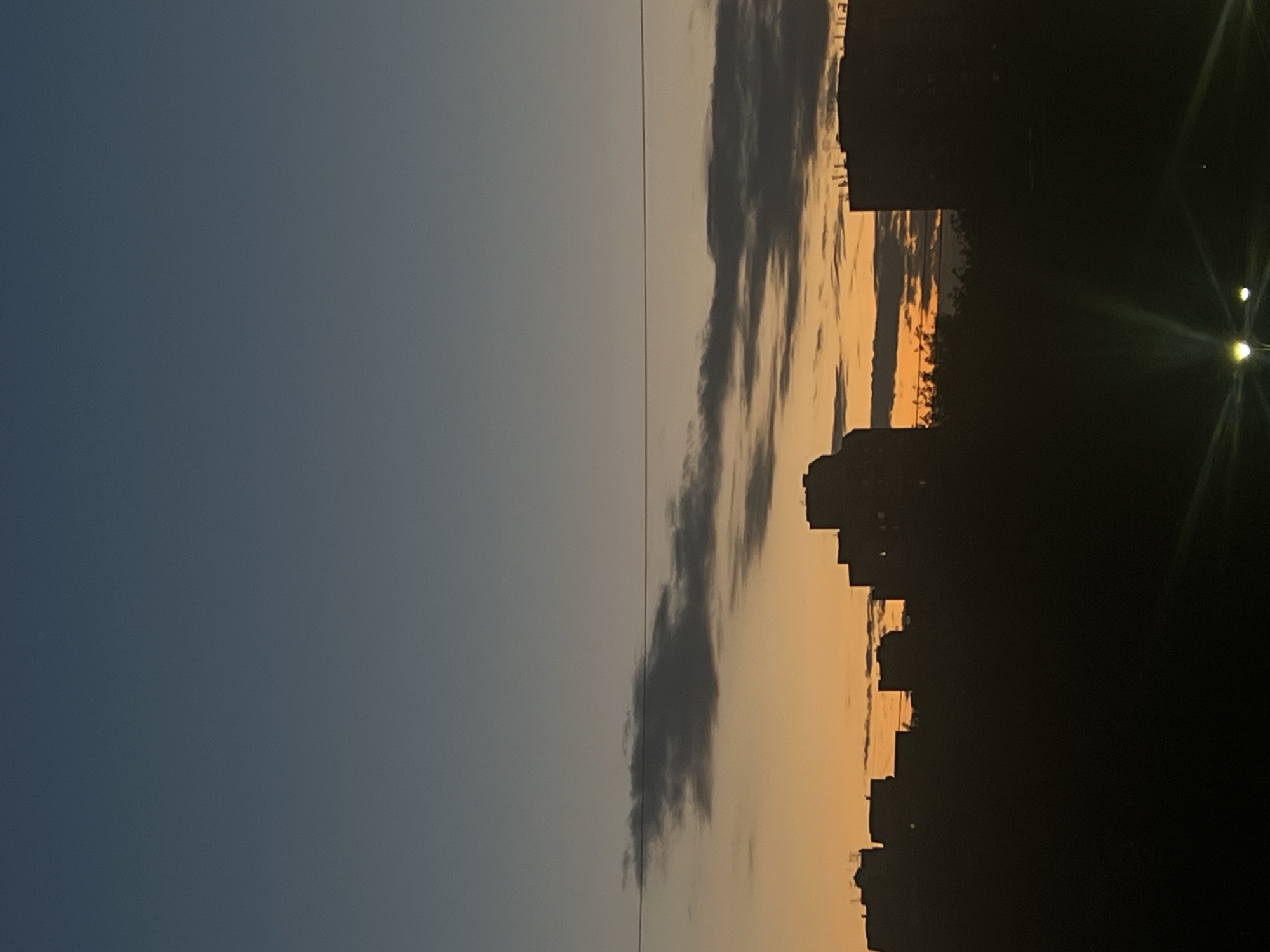 A cityscape at dusk is depicted, with the silhouettes of apartment buildings against an orange and blue gradient sky. The sky transitions from a deep blue at the top to a vibrant orange just above the horizon. Several dark clouds are scattered across the sky, with some appearing as thin, wispy lines and others as denser formations. A single power line stretches horizontally across the mid-section of the image. The lower portion of the image is dominated by the dark silhouettes of trees and buildings, obscuring the ground. Two streetlights are visible in the bottom right corner, emitting bright flares. The scene appears to be an urban environment in Saint Petersburg, Russia, characterized by the architecture of the buildings.