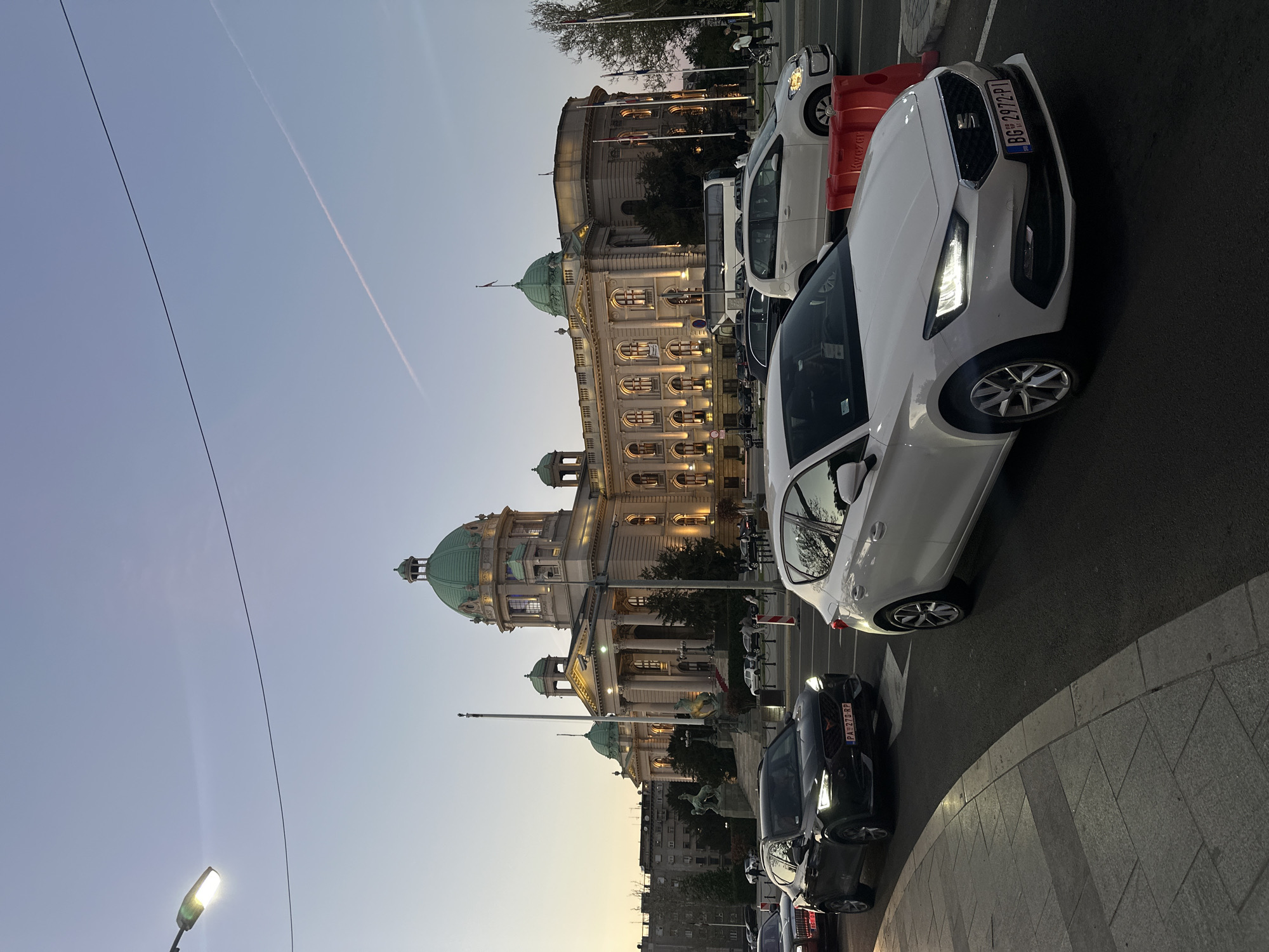 This image captures an evening street scene in Belgrade, Serbia, presented with a 90-degree counter-clockwise rotation. The central subject is the grand House of the National Assembly of Serbia, a classical building adorned with multiple green domes and illuminated windows, creating a warm glow against the fading daylight.
In the foreground, several vehicles are parked along a curved street. A white car, likely a SEAT Leon, is prominent, with its LED headlights on. Its Serbian license plate reads "BG 2972-PI," indicating registration in Belgrade. Behind it, a black car is visible with the license plate "PA 278RP," registered in Pančevo. Another white car and a partial view of a white bus are further down the road. A red plastic barrier or cone is situated beside the white car, with a partially visible word that appears to be "Krozen".
The time of day is clearly dusk or early evening, characterized by the light blue sky, which shows a faint contrail, and the active streetlights and building illumination. A power line spans across the upper portion of the sky. The scene is calm, depicting an urban setting at twilight. No people are visible.
