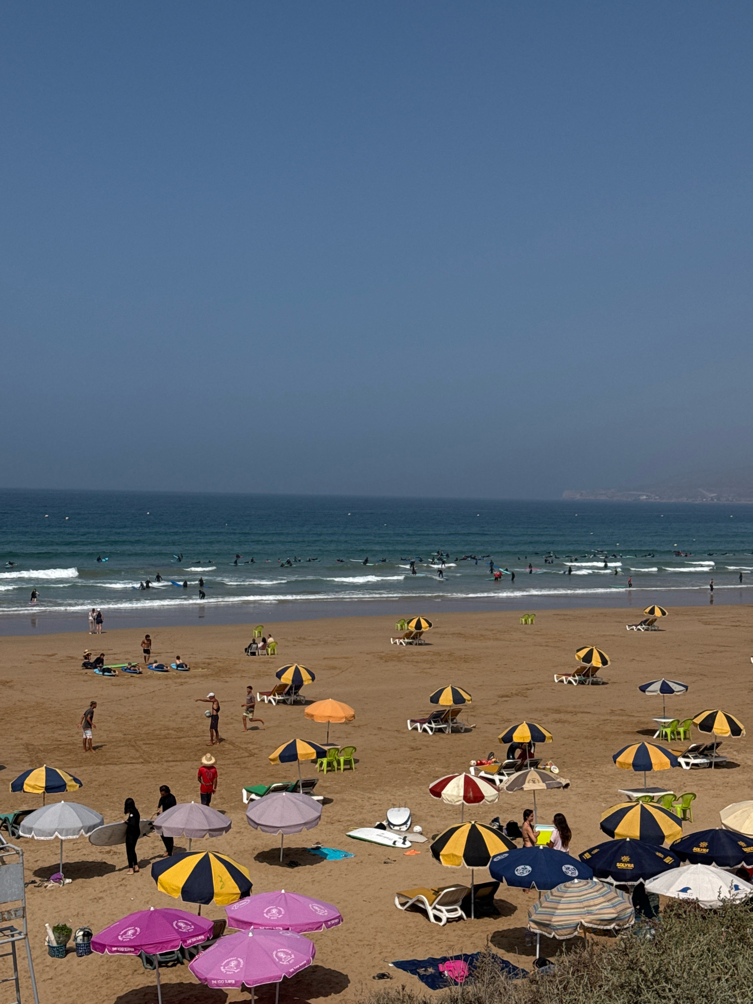 The image depicts a bustling beach scene in Aourir, Morocco, characterized by a wide expanse of golden sand meeting the blue ocean. The sky is a clear, light blue, with a hint of haze near the horizon where distant landforms are visible. The ocean is a vibrant turquoise, with white-capped waves rolling towards the shore, where numerous surfers are scattered, some actively riding waves and others waiting their turn.
The beach itself is populated with beachgoers enjoying the sun and surf. Loungers and umbrellas are strategically placed across the sand, offering shade and comfort. There are many colorful umbrellas, including yellow and black striped ones, purple ones, and blue and white checkered ones. People are seen relaxing on loungers, walking along the shore, and gathering under umbrellas. Several individuals are in the process of surfing or carrying surfboards.
In the foreground, the sand is textured and shows the footprints of those who have walked across it. The vibrant colors of the umbrellas and beach towels create a lively atmosphere. The scene suggests a typical summer day at a popular tourist destination, with activities centered around water sports and relaxation.