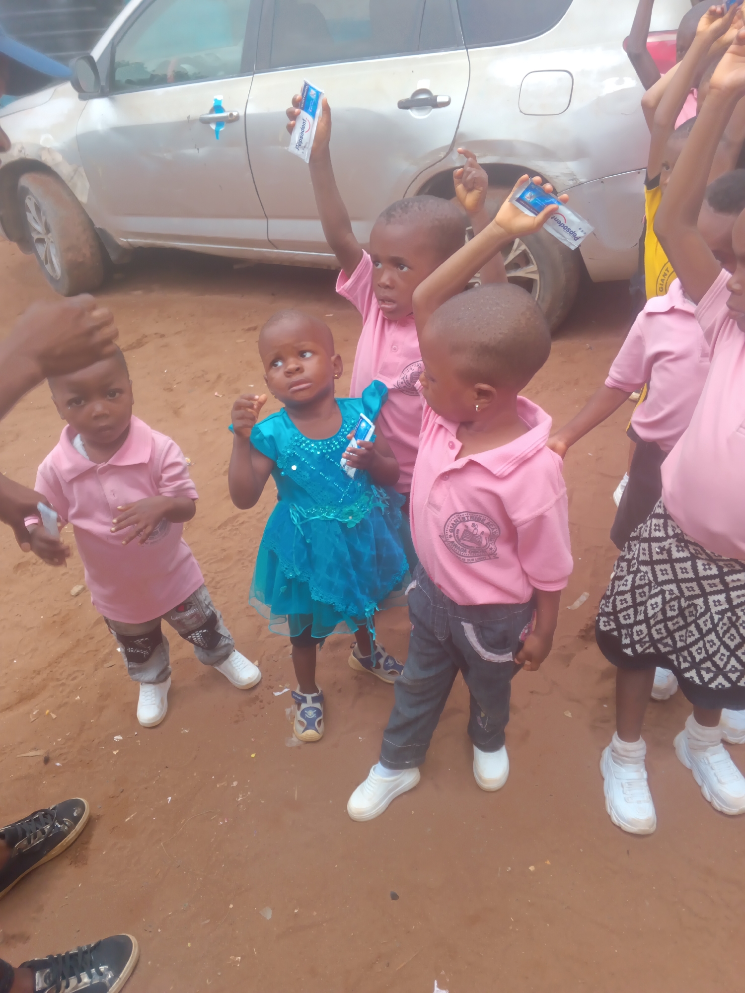 In Onitsha, Nigeria, a group of young children, likely students, are gathered outdoors. The ground is a dusty, reddish-brown earth, scattered with small white and yellow debris, possibly trash. A silver SUV is parked in the background, its right rear tire is muddy.
The children are dressed in casual clothing. Several boys wear pink polo shirts, some with school crests visible, paired with dark pants. One girl wears a bright turquoise dress. White sneakers are a common footwear choice among the children.
One child in the foreground holds a small, rectangular package, possibly toothpaste, with the brand name "Pepsodent" visible. Other children appear to be interacting, with hands raised and bodies angled towards each other. The overall scene suggests an informal outdoor gathering or activity, possibly related to a school event or distribution of items.