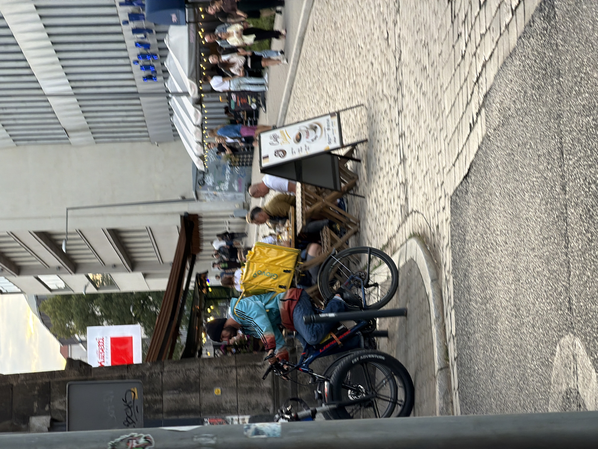 In an urban setting, a male food delivery person wearing a blue jacket and black beanie rides a dark blue electric bicycle with a large yellow delivery bag strapped to the rear rack. He is moving along a cobbled street in front of a building with a glass-fronted facade and blue signage. To the left of the delivery person, a dark gray pole stands, and a stone building with graffiti is visible. In the background, several people are seated at tables outside a cafe, with awnings providing shade. The street is paved with cobblestones, and there is a section of asphalt in the foreground. The sky above is overcast.