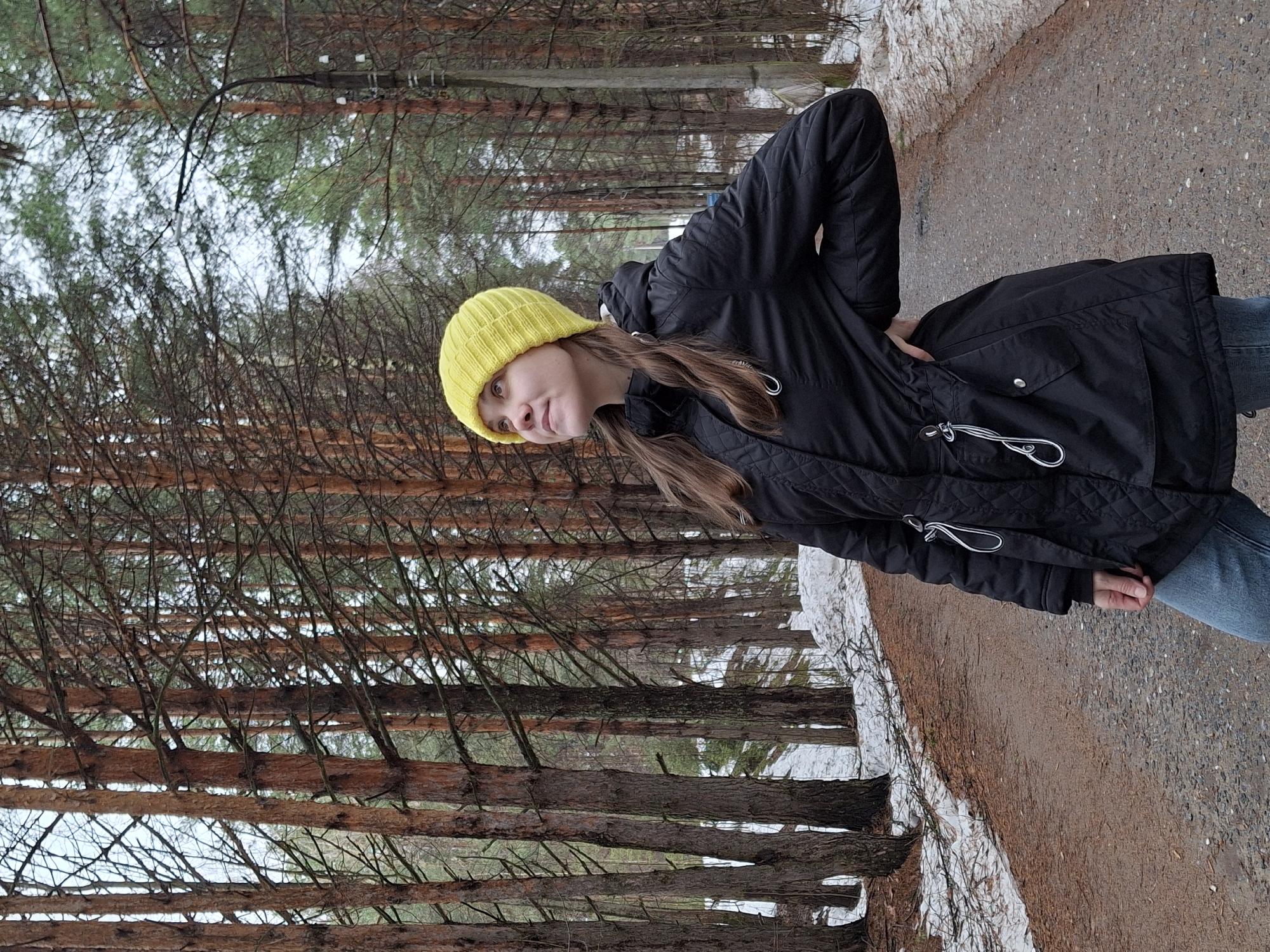 A young woman in a black quilted parka, blue jeans, and a bright yellow knit beanie is standing on a path in a forest. The path is made of gravel and dirt, with patches of snow visible along the edges. The forest is dense with tall, thin trees. The photo was taken in the city of Chaikovsky, Russia.