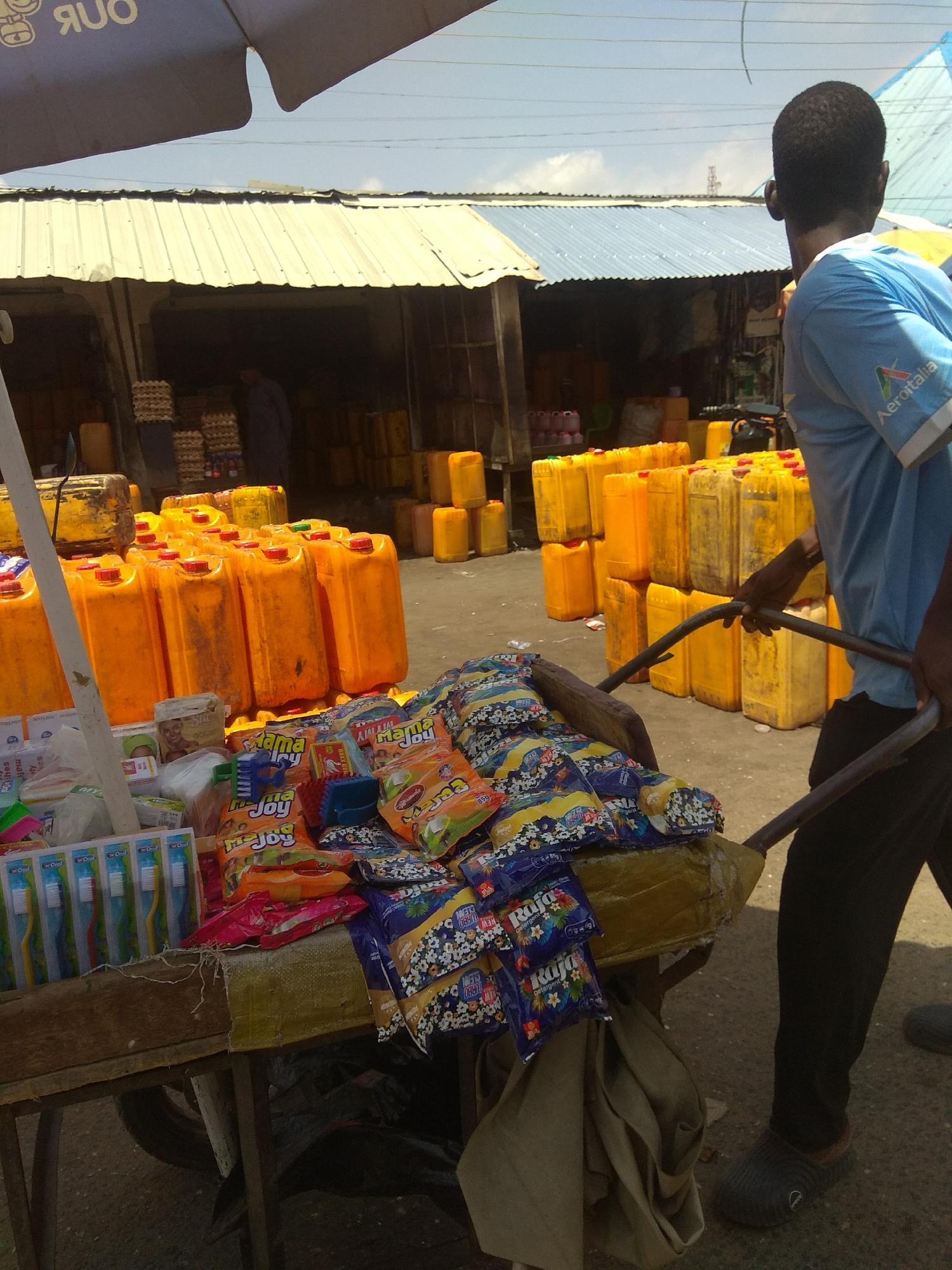The image depicts a street scene in Jalingo, Nigeria, likely a market area, with a focus on a vendor pushing a cart laden with goods. A man in a blue t-shirt with the "Aeroitalia" logo on the sleeve is pushing a cart from the right side of the frame. The cart is filled with various items, including packages of detergent and toothbrushes. Behind the cart, large yellow plastic containers, possibly for liquids, are stacked in rows. The environment appears to be outdoors with a roofed structure overhead, suggesting a covered market stall or an open-air stall with awnings. The ground is paved with asphalt or concrete. The overall impression is one of commercial activity in a Nigerian city.