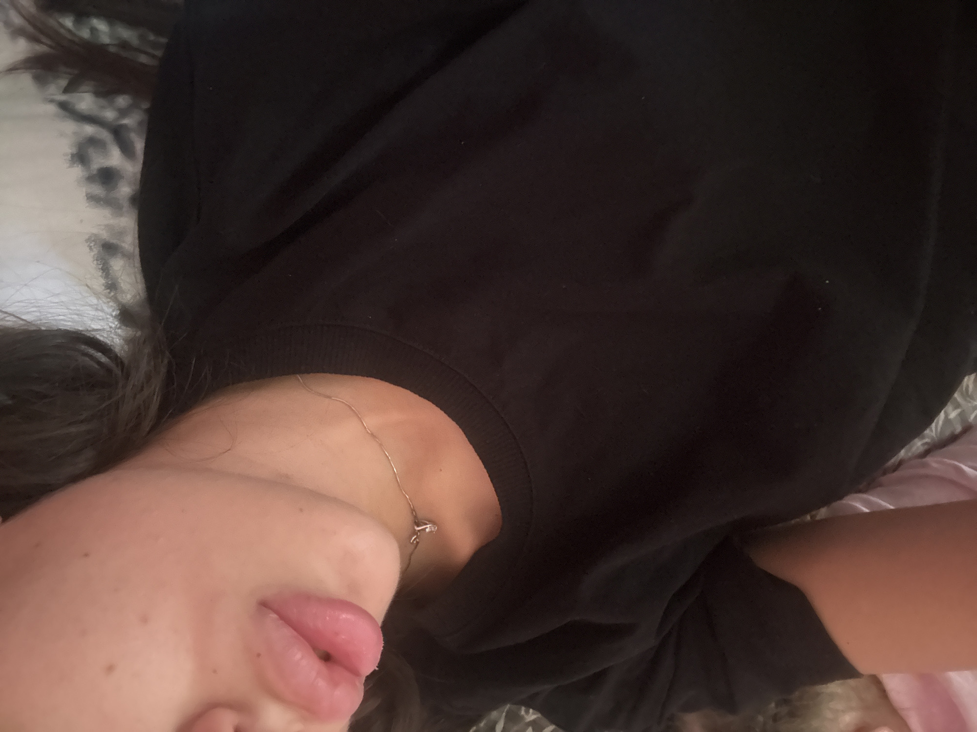 The image is a close-up, medium-shot, over-the-shoulder view of a person wearing a black t-shirt, lying down. The person's face is partially visible, showing their lips, chin, and neck. They are wearing a thin, silver necklace with a small, delicate pendant. The background is blurred and appears to be a bed with a patterned comforter. No specific location in Basarabeasca, Moldova can be identified from the visual information provided.