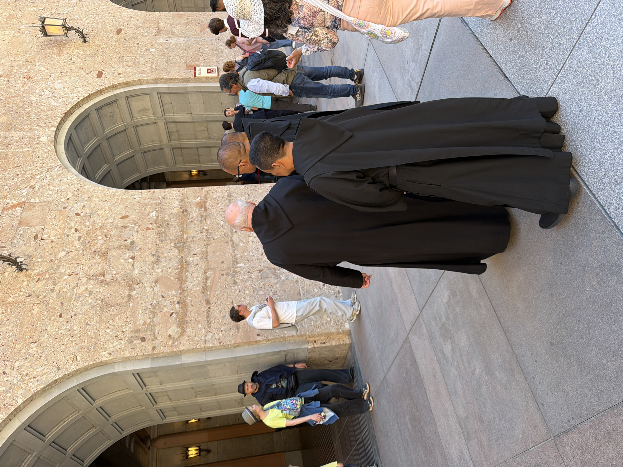 The image displays a bustling outdoor scene at what appears to be a historic or religious complex, likely a monastery given the context of Monistrol de Montserrat, Spain. In the foreground and central part of the frame, several men are prominently featured, dressed in long, dark, monastic-style robes. They are seen from behind, facing towards the right side of the image, appearing to walk in procession or a queue. Their heads are mostly bald or have very short hair.
Behind these robed figures, and further into the midground, a diverse group of visitors in casual clothing is visible. These individuals wear jeans, t-shirts, and other everyday attire, some with backpacks. One man wears a light blue shirt and jeans, while another is in a white t-shirt and light-colored pants. Near the bottom left, additional visitors are present, including one wearing a brimmed hat and a brightly colored shirt.
The setting is characterized by imposing stone architecture. A high wall, constructed from light-colored speckled or terrazzo-like stone, dominates the left side of the frame. Two large, deep arched doorways or passageways are carved into this wall, revealing intricately coffered ceilings within their recesses. A decorative black wrought-iron lantern is mounted on the stone wall near the top left corner. The ground is paved with large, grey square tiles.
The activity suggests a public gathering at a significant site, possibly a pilgrimage or a tourist visit, with some individuals apparently moving in an organized manner. The scene is well-lit by natural daylight, indicating clear weather, likely late morning or afternoon. A small, unreadable red sign is affixed to the stone wall between the arches.