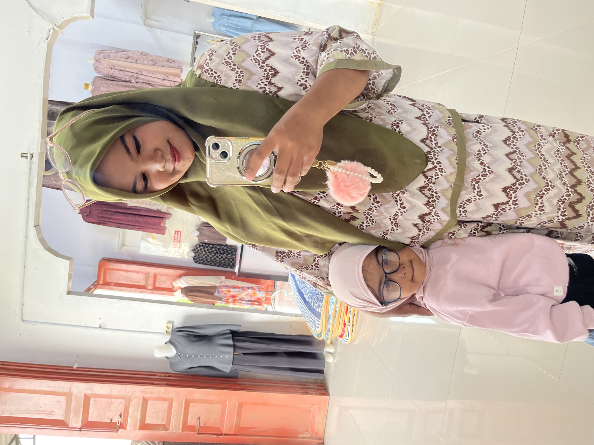 In a retail establishment in Blangmajron, Indonesia, an adult female wearing a green hijab and glasses is taking a selfie. She is holding a smartphone, which has a fluffy pink pom-pom keychain attached. Beside her, a young child, also wearing a pink hijab and glasses, is looking towards the camera. The background shows clothing racks with garments, mannequins displaying attire, and large red doors. The floor is tiled and reflective. The overall impression is of a mother and child posing for a photo inside a clothing store.