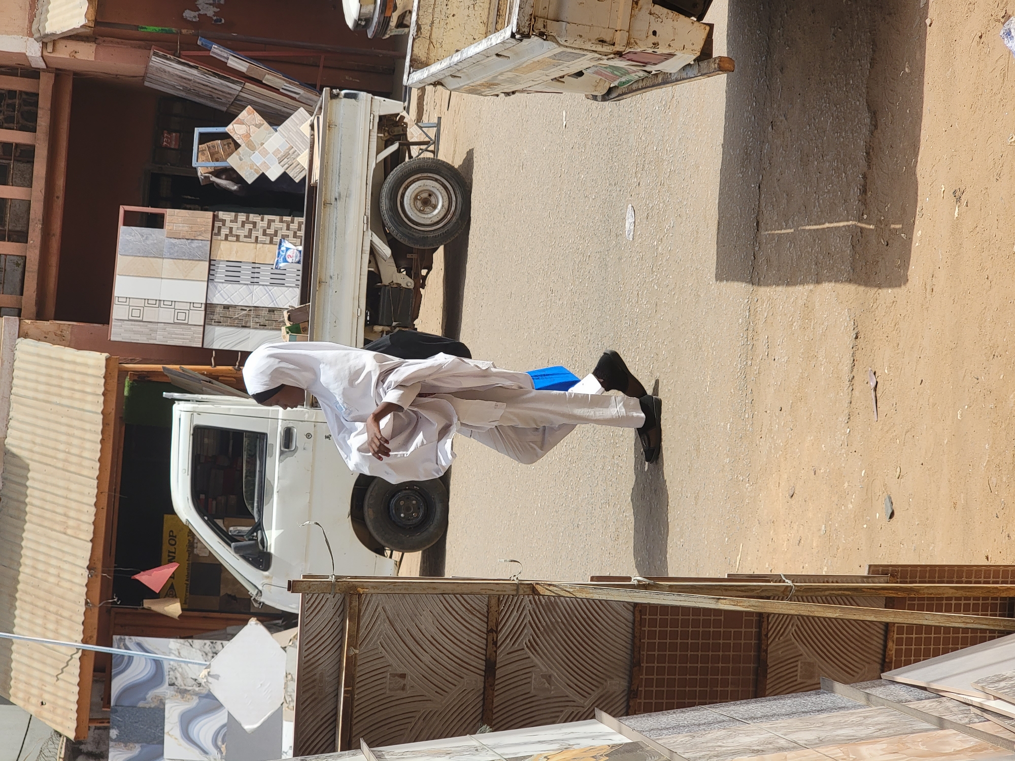 A man dressed in white stands on a street next to a white pickup truck loaded with tiles, possibly in Maiburiji, Nigeria. More tiles are stacked near a corrugated metal structure. The street is paved, and the environment suggests a commercial area.