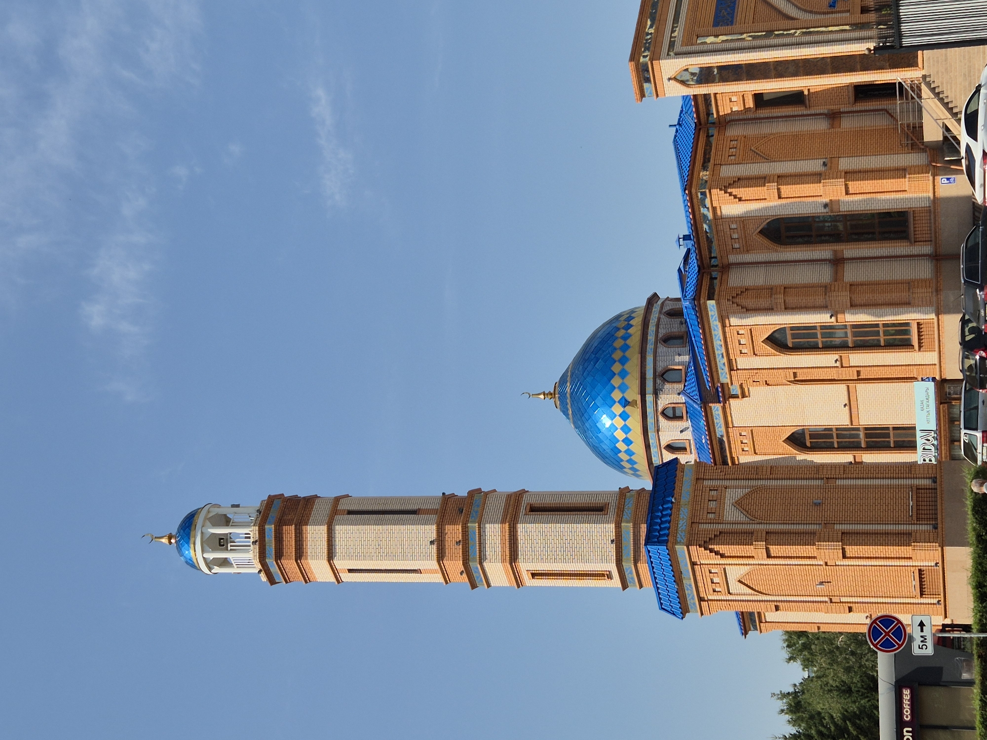 The image shows a bright orange brick mosque with a blue tiled dome and a tall minaret. The mosque is adorned with blue and gold decorative elements. The sky is clear blue with a few wispy clouds. In the foreground, there is a sidewalk and some parked cars. A sign with a red circle and a blue cross indicates no parking. Another sign shows "5M" with an arrow pointing to the right. Trees are visible in the background. The mosque is identified as the Nur-Astana Mosque in Kokshetau, Kazakhstan.