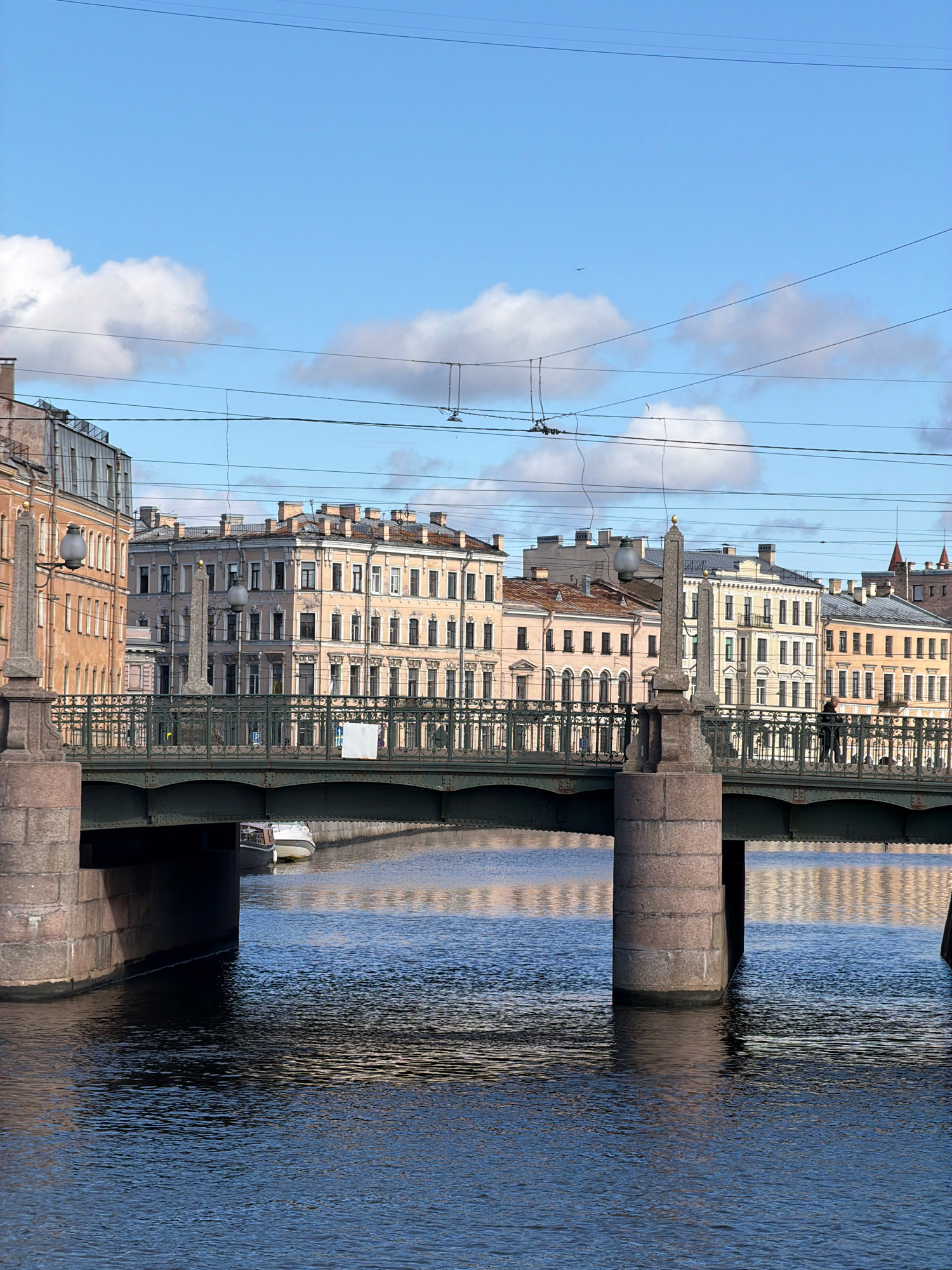 This is an eye-level, medium shot of a bridge crossing a canal in Saint Petersburg, Russia, on a clear day with scattered clouds. The bridge, made of metal with decorative railings, spans the canal. Its stone support pillars are visible on either side of the frame. On the bridge, two people are walking, one from the right and one from the left, neither interacting. A small white boat is moored under the bridge on the left side. The canal water is dark blue and reflects the sky and buildings. The background features several multi-story buildings with distinct architectural styles, typical of Saint Petersburg, including ornate windows and mansard roofs, painted in shades of beige and yellow. Power lines are visible crisscrossing the sky.