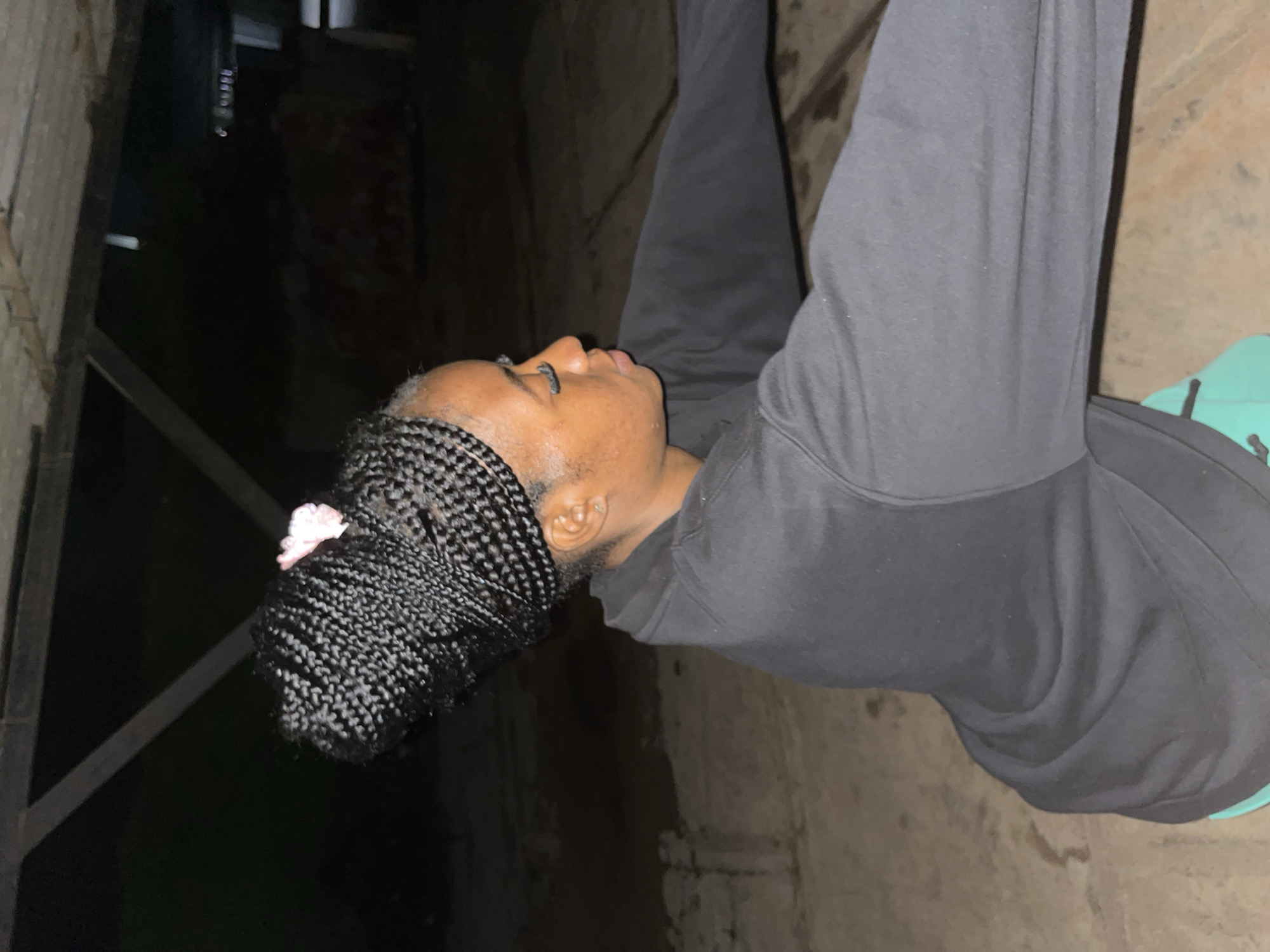 A young Black woman is depicted in a medium shot, turned to her right, with her face in profile. She has tightly braided hair styled in a large bun atop her head, adorned with a small pink accessory. Her complexion is medium-toned, and she wears false eyelashes. She is dressed in a black hooded sweatshirt, and a glimpse of turquoise athletic shorts is visible below. The background is dark and appears to be an outdoor area with a rough, concrete floor and overhead wooden beams, suggesting an unfinished or rustic setting. The lighting is dim, creating shadows and an overall low-light atmosphere. Based on common architectural styles and ambient conditions, the scene is likely located in Jos, Nigeria.