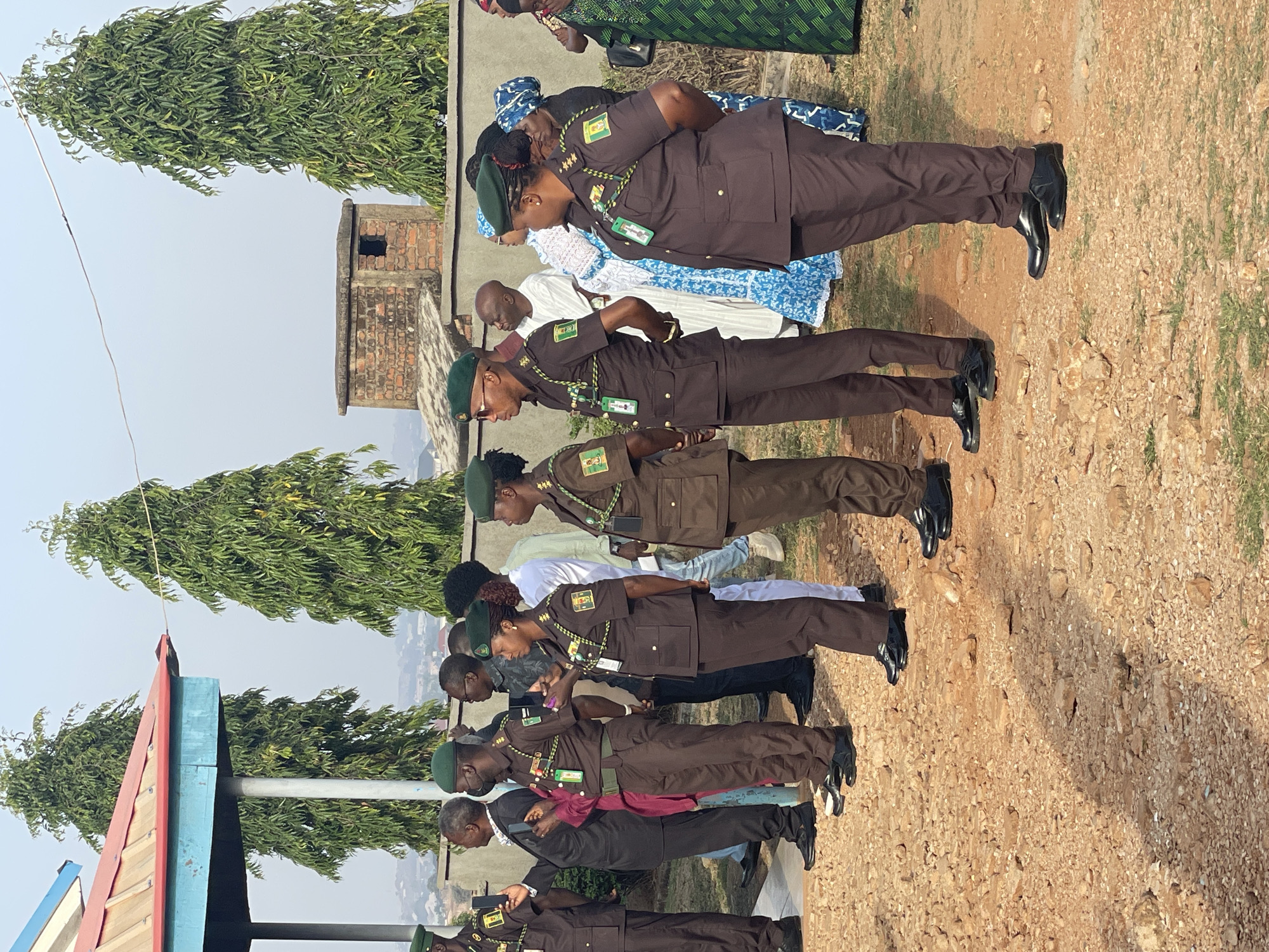This outdoor image depicts a formal gathering involving personnel from the Nigerian Correctional Service (NCS) and civilians in Jos, Nigeria, under bright daylight.
In the foreground and mid-ground, a line of uniformed individuals stands formally. They are dressed in dark brown uniforms with green berets and black polished shoes. Each uniformed person wears a round badge on their left chest, clearly displaying "NIGERIA CORRECTIONAL SERVICE" along with an emblem. Many also have ID cards attached to green lanyards, which distinctly show "NCS" at the top, confirming their affiliation. Both male and female personnel are visible within this uniformed group, standing attentively.
Behind the uniformed individuals, several civilians are observing the proceedings. Some are dressed in traditional African attire, including blue and white patterned dresses and a green patterned dress, while others wear everyday clothing.
The setting is an unpaved, dusty area with light brown soil and scattered small rocks, suggesting an open field or ceremonial ground. Tall green trees with dense foliage are prominent in the background and along the sides. A small, weathered brick structure with a dark window is visible behind the people. On the far left, a portion of a light-colored structure with blue and red painted elements can be seen, possibly a part of a stand or shelter. The sky is clear and bright, indicating sunny weather, likely mid-morning to early afternoon given the visible shadows. The scene conveys a sense of formality and official observance.