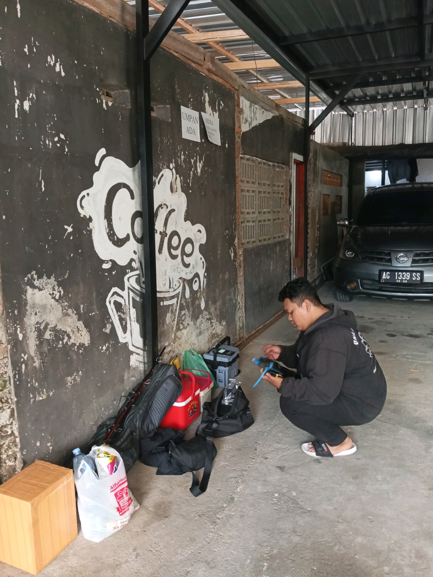 In Prigi, Indonesia, a young man in a black hoodie and pants crouches on a concrete floor. He holds a blue and yellow object, appearing to be assembling or repairing something. Next to him, on the left, are several bags, including a red cooler, a fishing rod case, and a white plastic bag from Alfamart containing a water bottle and other items. A brown cardboard box sits further to the left. Behind the man, a dark grey Nissan sedan with license plate AG 1339 SS is parked. The background features a dark, textured wall with white graffiti depicting a coffee cup and steam, and a corrugated metal roof structure. The environment appears to be an outdoor covered area, possibly a garage or workshop.
