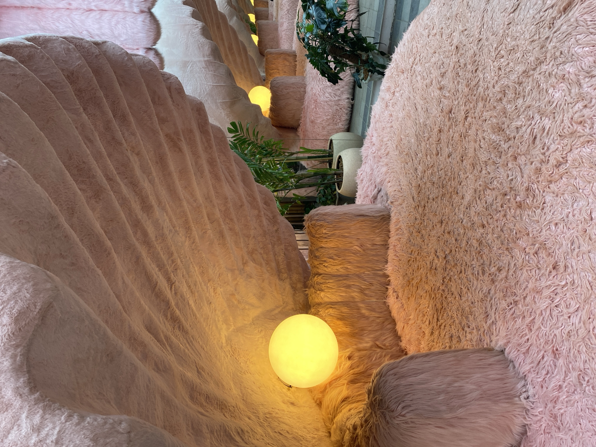 This media file captures an interior scene with a soft, textured aesthetic in Pattaya City, Thailand. The dominant subjects are abstract, undulating forms covered in plush, pink, and peachy faux fur. These forms create a unique architectural or decorative element, resembling oversized waves or dunes. Scattered throughout the scene are spherical lamps, emitting a warm, yellow glow that illuminates the textured surfaces.
The setting appears to be an indoor space, possibly a lounge area or a unique retail environment, characterized by its soft lighting and tactile surfaces. The faux fur covering extends across various levels and shapes, creating an immersive and cozy atmosphere. In the background, glimpses of lush green plants in light-colored planters and what appear to be striped vertical elements suggest a contemporary interior design.
There are no people visible in the media. The activity taking place is passive; the scene is static, focusing on the visual and textural qualities of the environment. The time of day is not definitively ascertainable, but the warm lighting suggests an indoor setting designed for evening or ambient illumination. The overall emotion evoked is one of comfort, whimsy, and perhaps a touch of surrealism due to the unusual materials and forms. No visible text is present in the image.