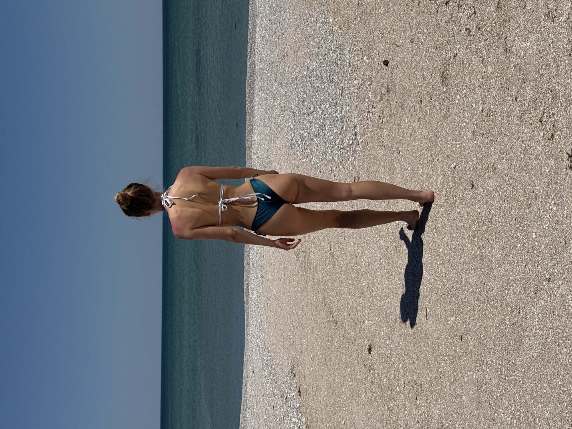 A person stands on a beach in Мысовское сельское поселение, Ukraine. The individual, seen from the rear, wears a white bikini top and dark bottoms. The sea is to their left, and a sandy, shell-covered beach stretches to the right. The person's shadow is cast on the sand. The sky is clear.