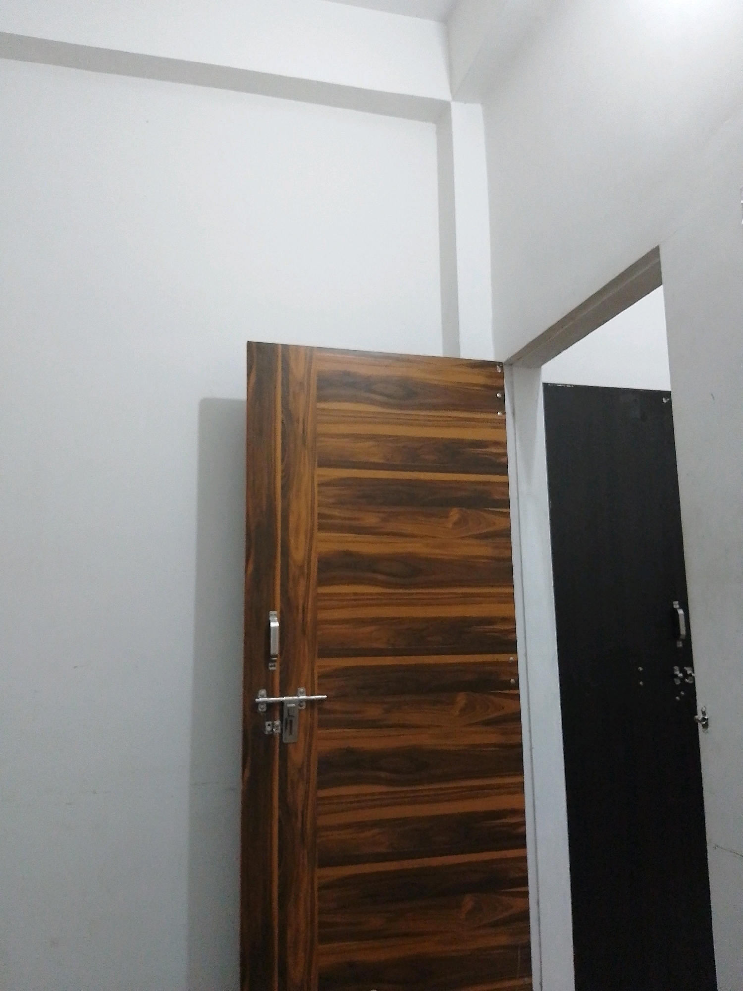 The image shows an interior view of a room with two doors. One door, on the left, is made of wood with a prominent grain pattern and has a silver handle and latch. The other door, on the right and partially visible, is solid black with a silver handle. Both doors are set within a white door frame. The walls of the room are painted white, and there is a white beam or trim along the ceiling. The scene appears to be a residential or institutional setting. The overall lighting suggests an indoor environment, possibly a hallway or entrance. There is no indication of the location being specifically in Jat Colony, India, as this is an assumption not supported by visual evidence in the image.