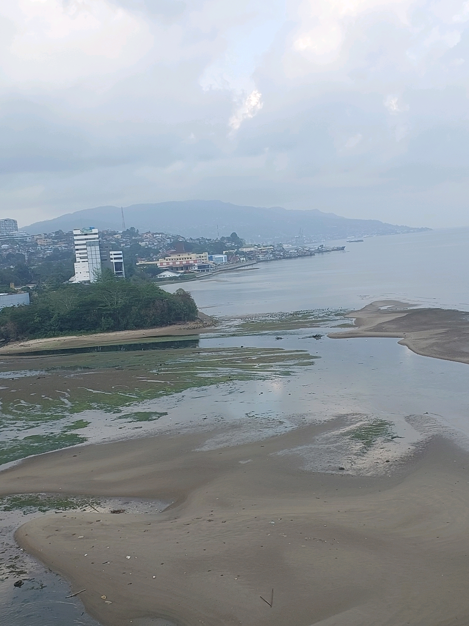 The image shows a panoramic view of Ambon Bay in Ambon, Indonesia. In the foreground, a mudflat is exposed, with patches of green algae and sandbanks. The water is calm and reflects the overcast sky. In the mid-ground, there is a small, tree-covered island. Behind the island, the city of Ambon stretches along the coastline, characterized by low-rise buildings and a few taller structures, including a prominent white and grey modern building. Further in the background, a range of hills forms a backdrop to the city. Several boats and a larger ship are visible in the bay. The overall atmosphere is subdued due to the cloudy weather.